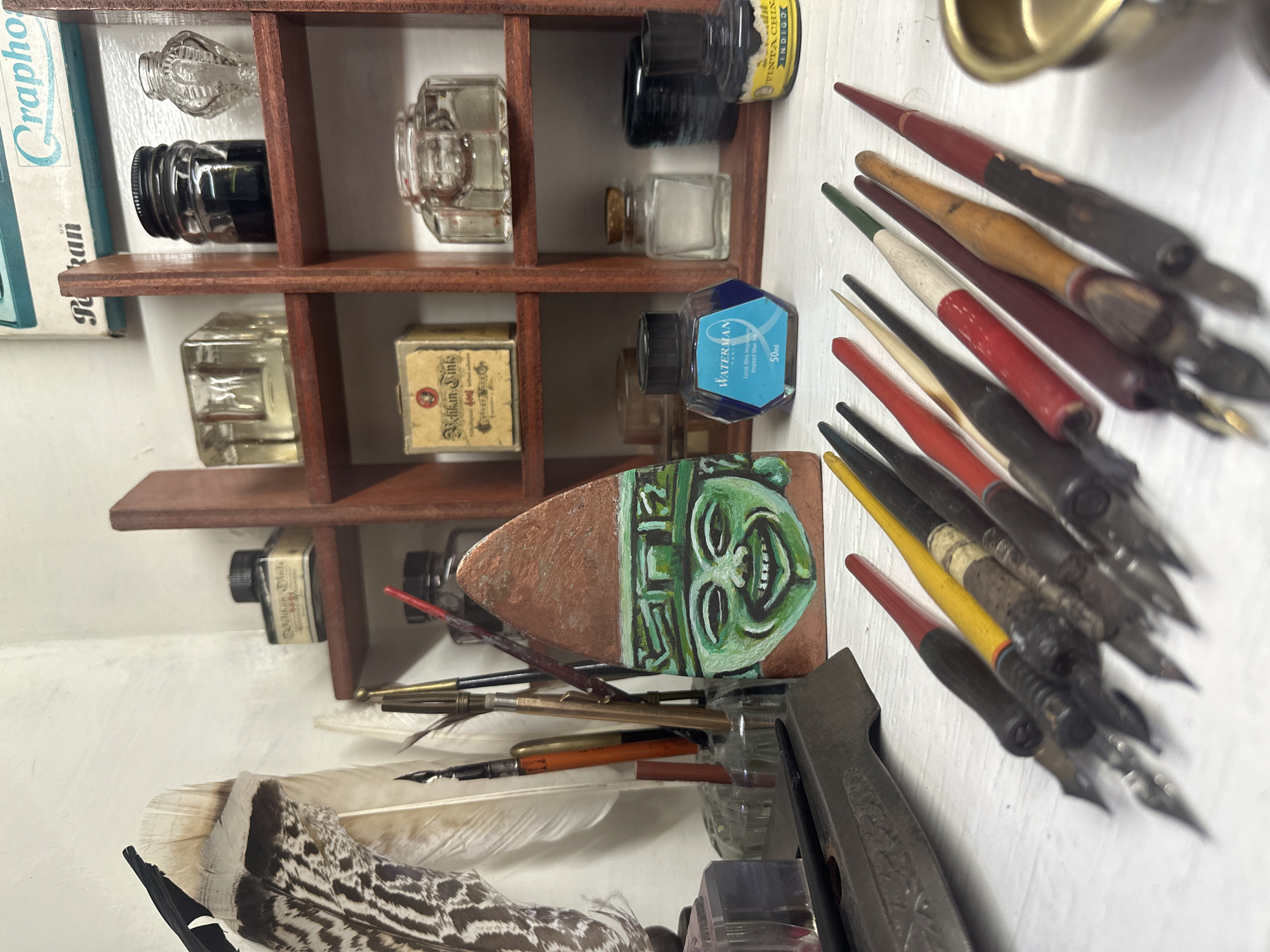 The image depicts a wooden shelf unit filled with various items related to writing and art. On the shelves are several bottles of ink, including one labeled "WATERMAN Encre Bleu Inspiration Inspired Blue 50ml" and another that reads "TINTA CHIN INDIGO." There is also a box that is labeled “Pelikan Sinke 4001” as well as “Pelikan Grapho.” The shelf unit holds a collection of antique inkwells and glass bottles.
Below the shelf unit, there's an assortment of dip pens, some with colored handles (red, yellow, and black). There's a feather with a black and white pattern lying near the pens. Also, there is a carved sculpture of a face that is green in color with the background appearing to be copper. The scene appears to be a workspace or a collection display. There is no indication of location in Mexico City, Mexico.