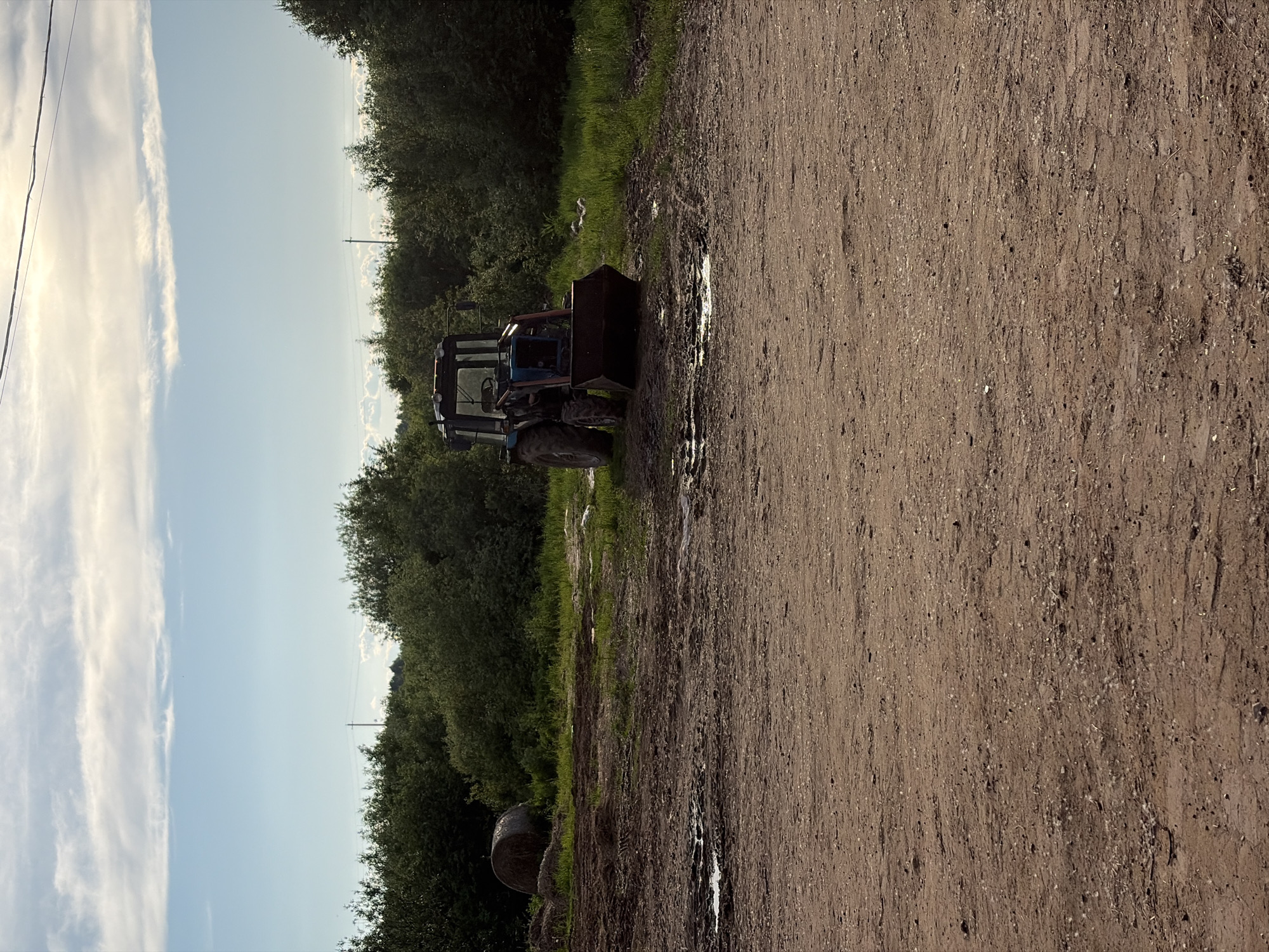 The image, when viewed in a corrected landscape orientation, displays a rural scene in Чумна, Russia, under a bright, partly cloudy sky. A wide, well-trodden dirt track, marked with puddles and reflections, dominates the foreground and extends towards the right side of the frame.
To the left of the dirt track, a narrow strip of green grass separates it from a dense line of dark green trees and bushes that forms a natural barrier in the middle ground. A blue tractor, appearing to be an older model, is parked stationary on the grassy edge of the track, positioned on the left side of the image. It is equipped with a front-end loader bucket. Further back and to the left, partially hidden by the grass and foliage, is a round bale of hay or silage.
The sky above features streaky white clouds on the left, transitioning to a clearer blue expanse on the right. Overhead power or utility lines are visible against the bright sky on the far left, extending across the upper portion of the scene. In the distant right, a utility pole stands vertically, silhouetted against the horizon line where the trees meet the sky. The lighting suggests it is late morning or afternoon. No people are visible, and the scene conveys a sense of stillness. There is no visible text.