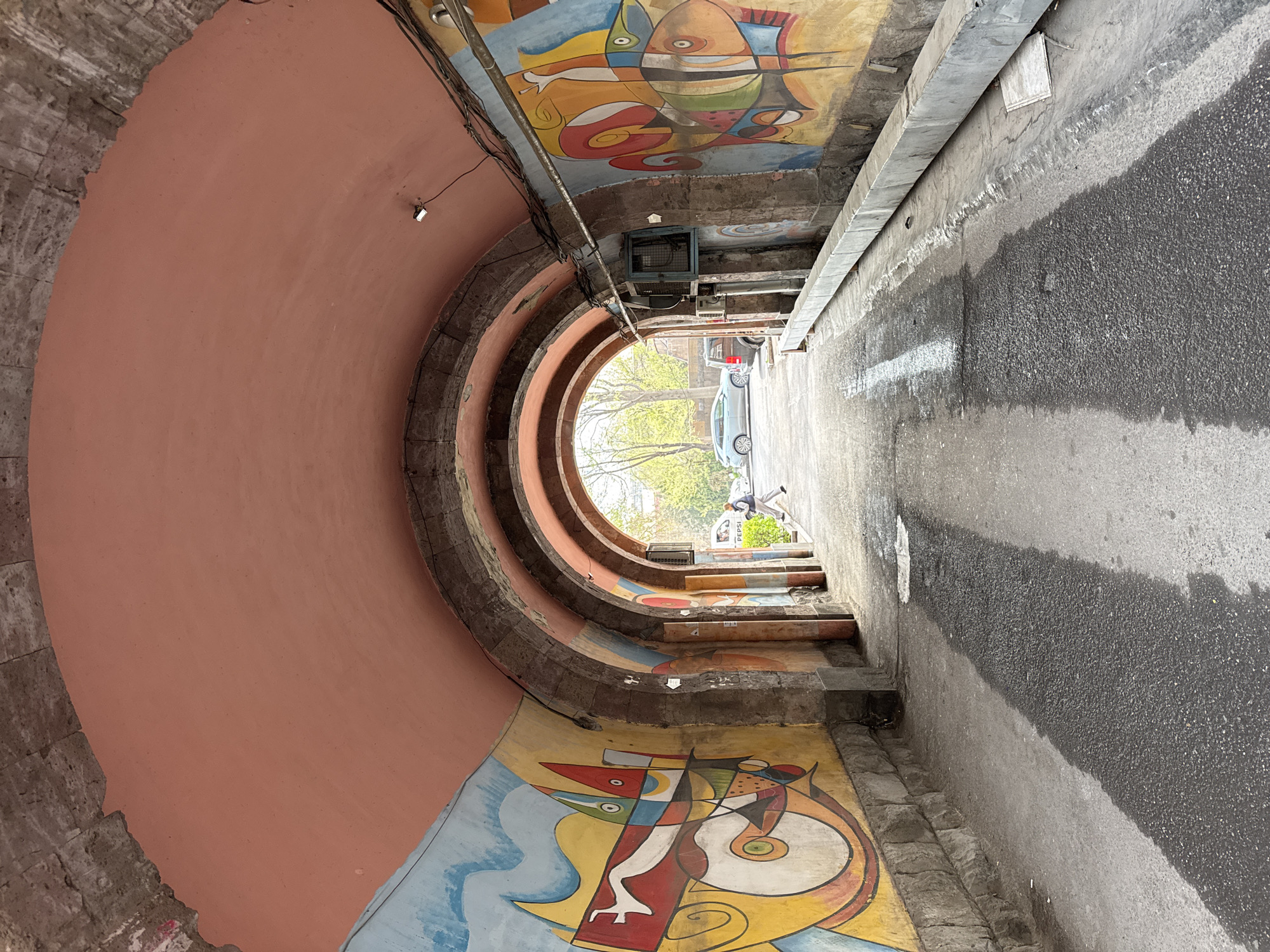 View looking down the Paparazzi Steps in Yerevan, Armenia. The stairwell is painted with colorful abstract murals. There is a car parked near the top of the steps, and a pedestrian walking away from the camera at the top. The exterior wall is pink, and there is an asphalt surface to the right of the stairs.