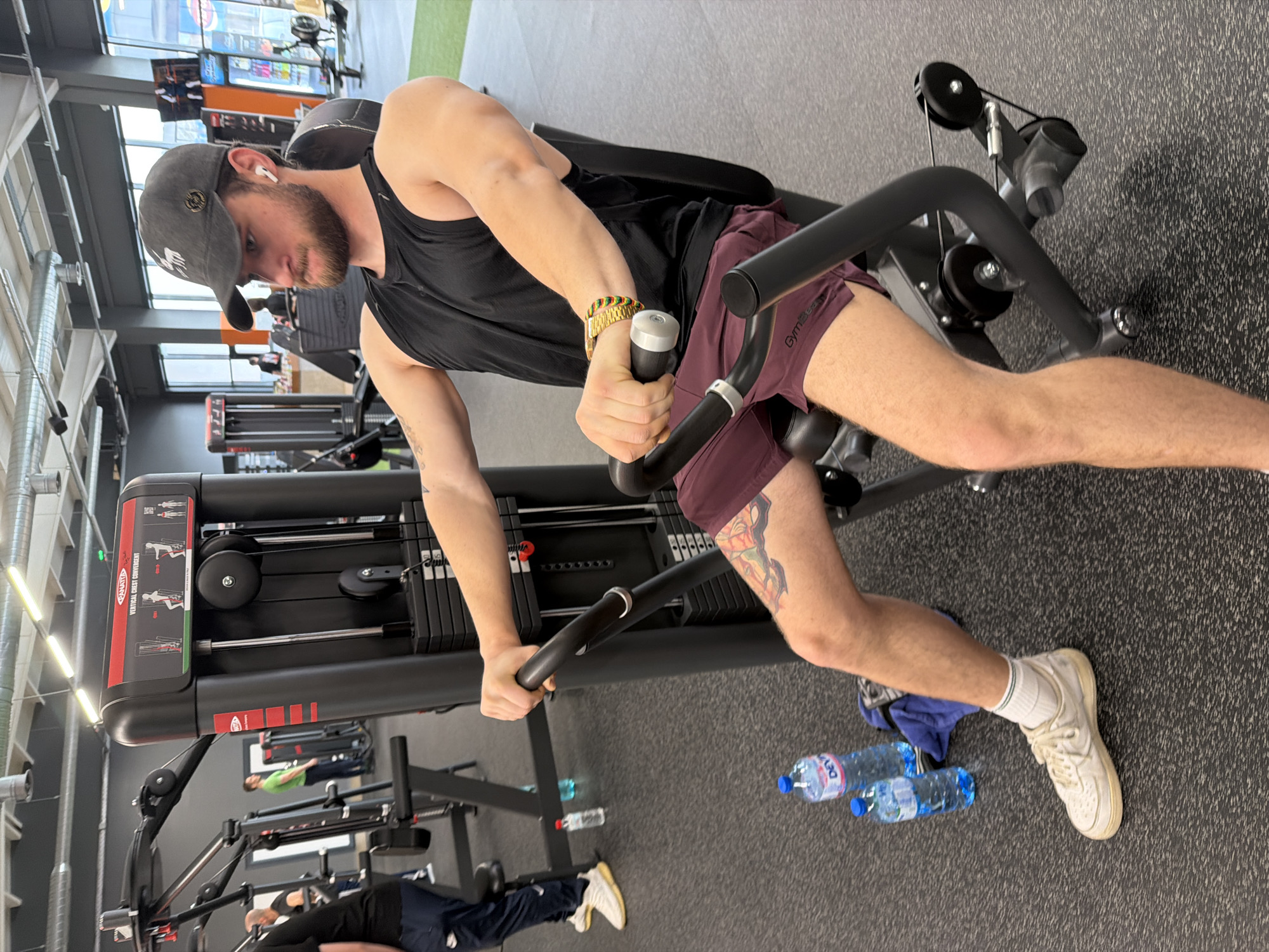 The image depicts a man in a gym, working out on a seated chest press machine. He is wearing a black tank top, maroon shorts, a grey baseball cap, and white sneakers. A gold watch and a bracelet adorn his left wrist. A tattoo is visible on his left thigh. Two water bottles and a blue towel sit on the floor next to his feet. Other gym equipment and people are visible in the background. The gym is located in Plovdiv, Bulgaria.