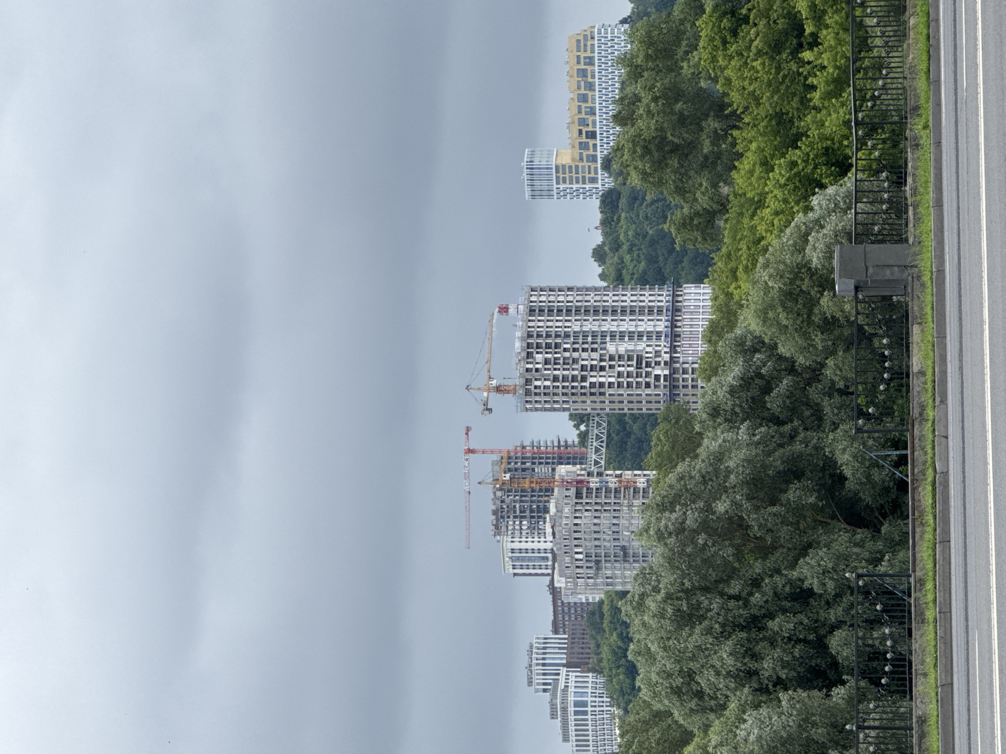 The image depicts a cityscape under a cloudy sky, with a focus on several multi-story buildings, some of which are under construction. Cranes are visible, indicating ongoing development. The foreground features a lush green area with mature trees, a metal fence, and a section of road. In the background, several modern apartment complexes and office buildings rise above the tree line. One building, a distinct yellow and white structure, is particularly noticeable. The overall impression is of a developing urban area surrounded by natural greenery. This scene is identified as being in Nizhny Novgorod, Russia.