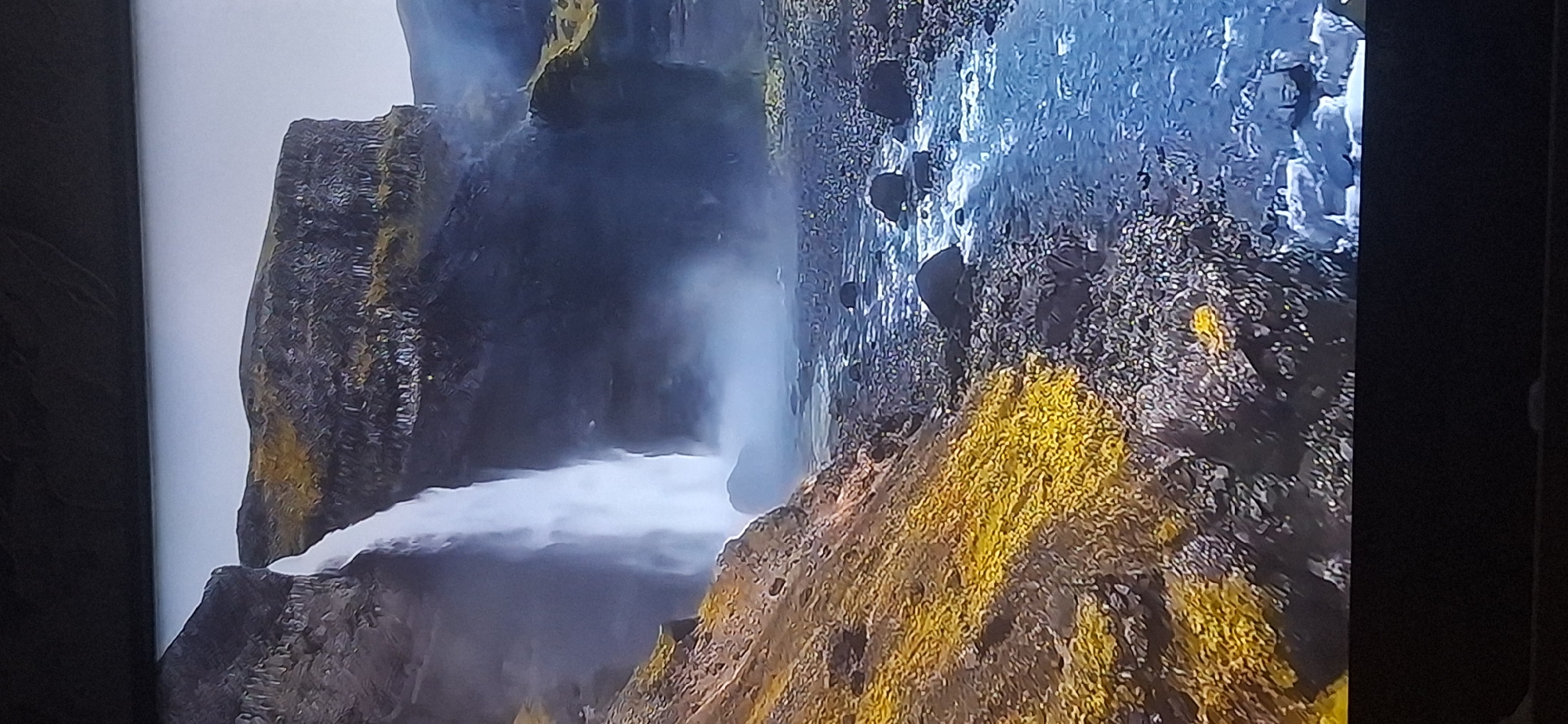 This is a high-angle, vertical, medium shot of a waterfall cascading down a rocky cliff. The waterfall is white and misty, with the mist obscuring the base of the falls and the surrounding rocky terrain. To the left of the waterfall, a steep, rocky slope is covered with patches of vibrant yellow vegetation. The foreground is dominated by a rocky riverbed with clear, blue water flowing between dark, rounded stones. The background features a sheer, dark cliff face with textured, columnar rock formations, partially covered in similar yellow vegetation. The sky above is a pale, overcast gray. The image does not depict any people or recognizable landmarks of Bila Tserkva, Ukraine.
