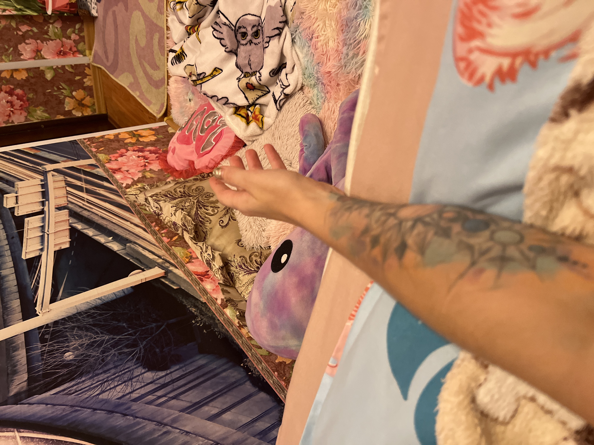 The image captures a close-up view of a person's arm with tattoos resting on a soft, multi-colored blanket. In the background, a purple stuffed animal with a black eye is visible, along with a pink heart-shaped pillow and a blanket featuring cartoon owls. The environment appears to be a bedroom, with floral wallpaper and a rug. The scene does not contain any elements that indicate it is located in Krasnoe-na-Volge, Russia.