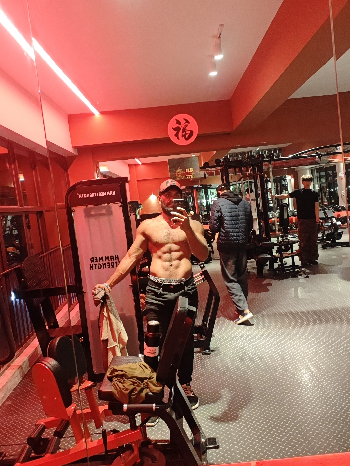 An interior image depicts a gymnasium within the Chongqing High-tech Zone, China. The environment features a gray diamond plate patterned floor and walls accented with red paint and red LED strip lighting. A prominent red circular plaque displaying the Chinese character "福" (fú) is mounted on a wall.
In the foreground, a shirtless, muscular male with a beard and a dark cap is taking a mirror selfie with a smartphone, obscuring his face. He wears dark trousers and sneakers, and holds a light-colored towel in his left hand, which rests on a "HAMMER STRENGTH" leg exercise machine. A black water bottle is attached to the machine's frame, and an olive-green garment is crumpled on its seat.
In the mid-ground, a second male, viewed from behind, walks from right to left, wearing a dark puffer jacket, dark trousers, and light sneakers. To the right, in the background, a third male stands with his back to the camera and arms slightly outstretched, dressed in a dark long-sleeved shirt and dark patterned trousers. Various other gym equipment is visible throughout the reflections and directly in the background.