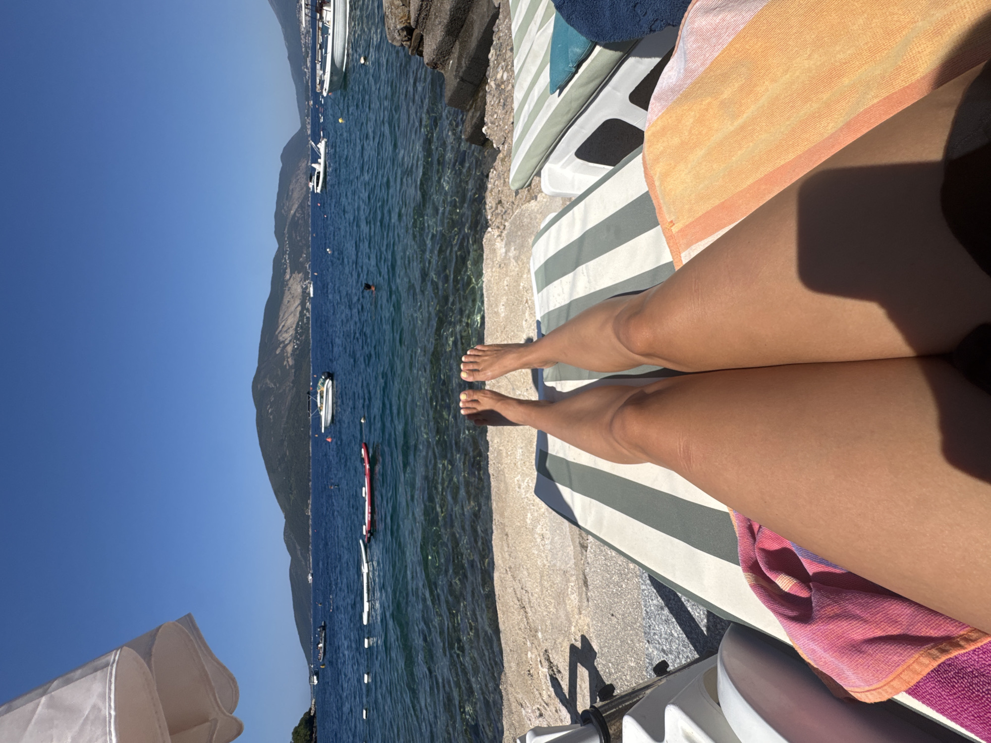A first-person perspective shot shows a person's legs and feet resting on a striped lounge chair on a beach. The legs are tanned and bare, with toenails painted yellow. The lounge chair has green and white stripes. A pink and orange striped towel is draped over the left side of the lounge chair, and a bright orange towel is on the right. In the background, the clear blue water of the sea is visible, with several boats and a person swimming. Mountains rise in the distance, and the sky is a clear blue. The scene is one of relaxation and a sunny day at the beach.