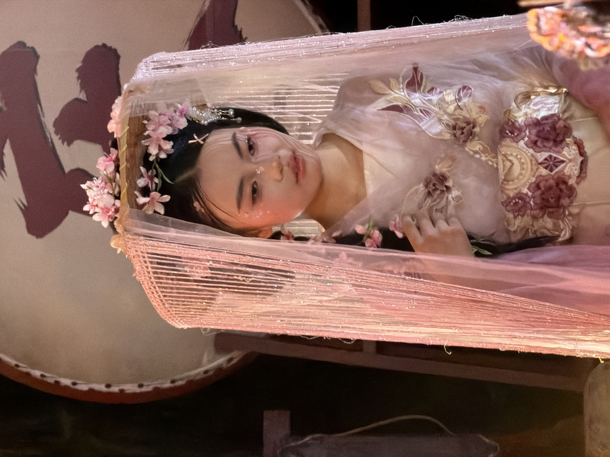 An East Asian female in a traditional costume and headdress adorned with pink flowers stands in front of a large, round object with Chinese calligraphy. She wears a veil with beaded fringe. The costume is sheer pink and embroidered with floral motifs. The background is dimly lit, with the large round object appearing to be a drum or screen. To the left, a wooden stand is visible. The scene appears to be set in Yuzhong District, China.