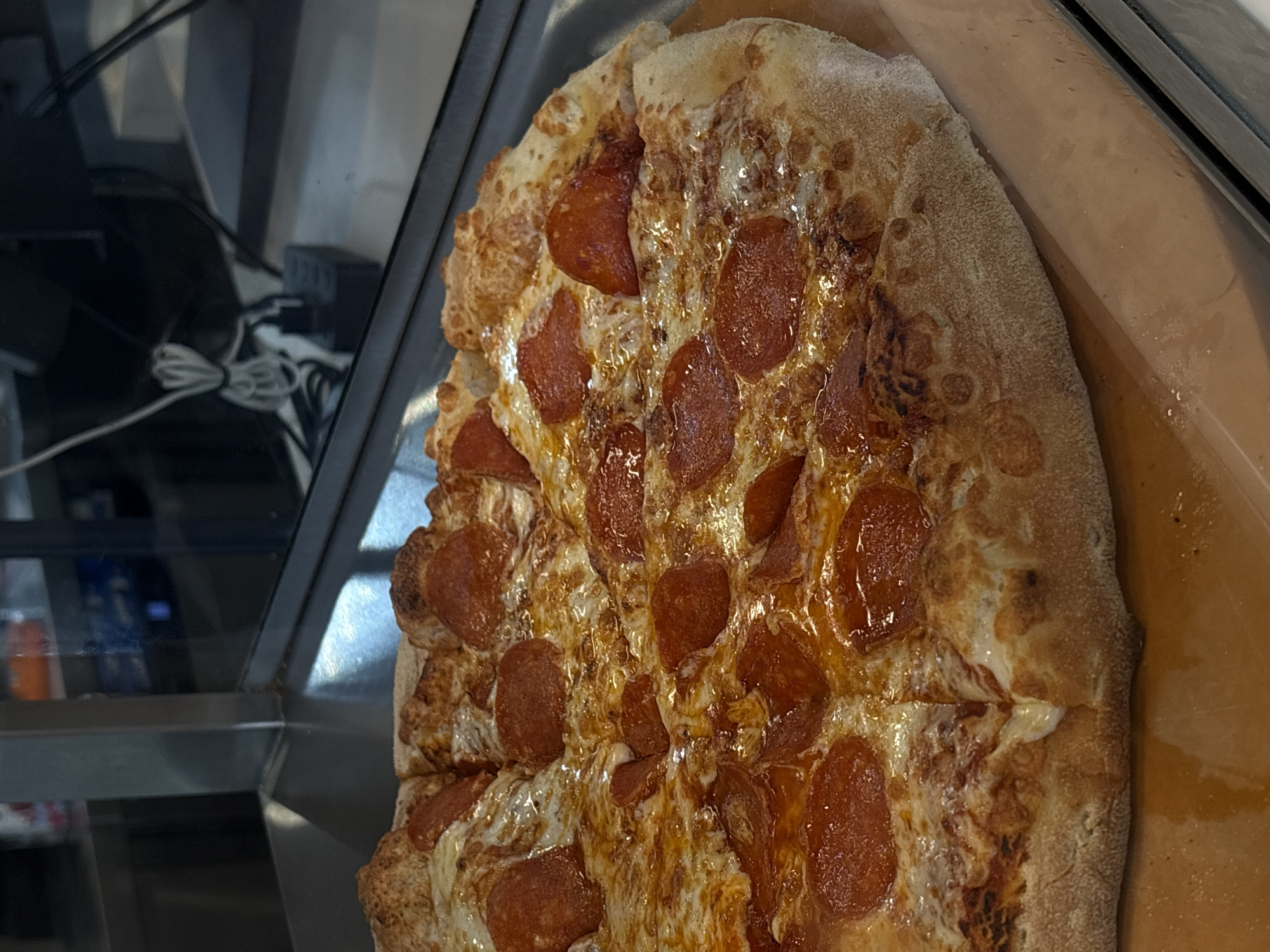 The image shows a partially cut pepperoni pizza. The pizza is topped with melted cheese and pepperoni slices. It is resting on a metal surface next to a glass panel where some wires can be seen. The location in the city of Birobidzhan, Russia cannot be determined from the image alone.