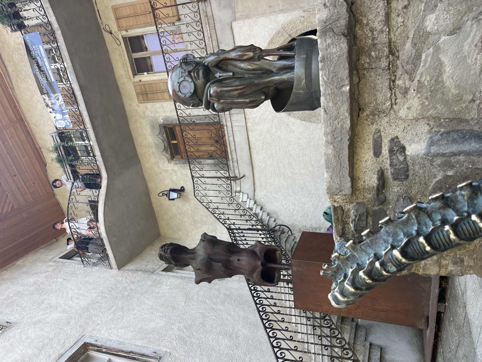 This image depicts an outdoor courtyard scene in Epagny, Switzerland, showcasing several sculptures and architectural elements. In the foreground, a bronze sculpture of a figure with large goggles and a diving helmet sits hunched over in a cylindrical base. To the left and slightly behind, another bronze torso sculpture stands on a rusted metal pedestal, with its head tilted upwards. Partially visible in the extreme foreground is a segment of what appears to be another sculpture, resembling a segmented, metallic appendage with brass accents.
In the background, a building with textured, cream-colored walls is visible. A balcony with ornate metal railings extends from the second floor, with several individuals standing on it. A blue banner with text, including "PHOTOGRAPHS HR GIGER PART II 1966-1975," is attached to the balcony railing. A set of stone stairs with a black metal railing leads up the side of the building towards the balcony. Wooden shutters are visible on the windows of the building. The overall impression is a courtyard with artistic installations set against a historic building.