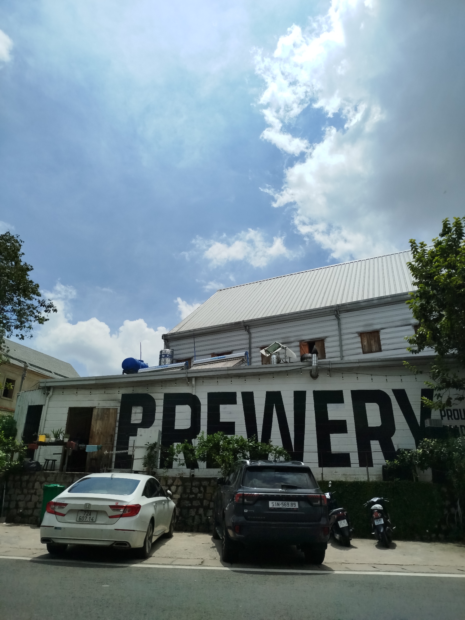 The image shows a bright, sunny day in Da Lat, Vietnam, with a clear blue sky and scattered white clouds. The main focus is a building with "BREWERY" painted in large black letters across its facade. The building appears to be made of light-colored horizontal planks. To the left of the building, there is a white sedan and to the right, a dark SUV and two motorcycles are parked. Lush green trees and bushes surround the building and parking area, suggesting a pleasant outdoor setting. The overall impression is one of a commercial establishment in a well-maintained urban environment.