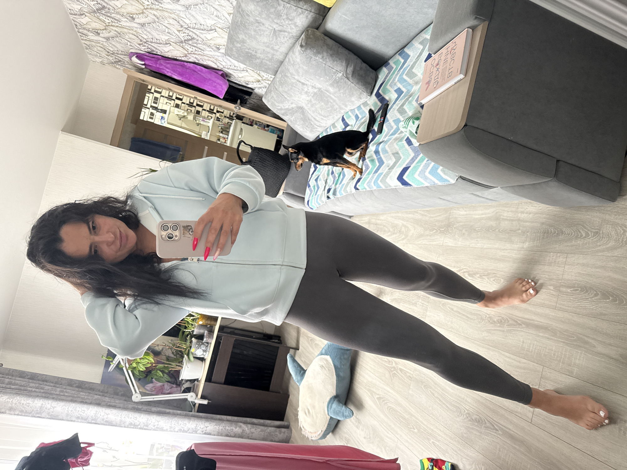 A woman is taking a selfie in an interior setting. She is wearing a light blue zip-up jacket and dark gray leggings, and her feet are bare. She holds a phone with a pink case in her right hand.
In the background, there is a gray sectional sofa with a small black and tan dog on it. A book is placed on the sofa armrest. Other items include a shark-shaped plush toy, houseplants, a black bag, luggage, and a shelf with various items. The walls are decorated with patterned wallpaper. The location is indicated as Kauchuk Stantsiya, Russia.
