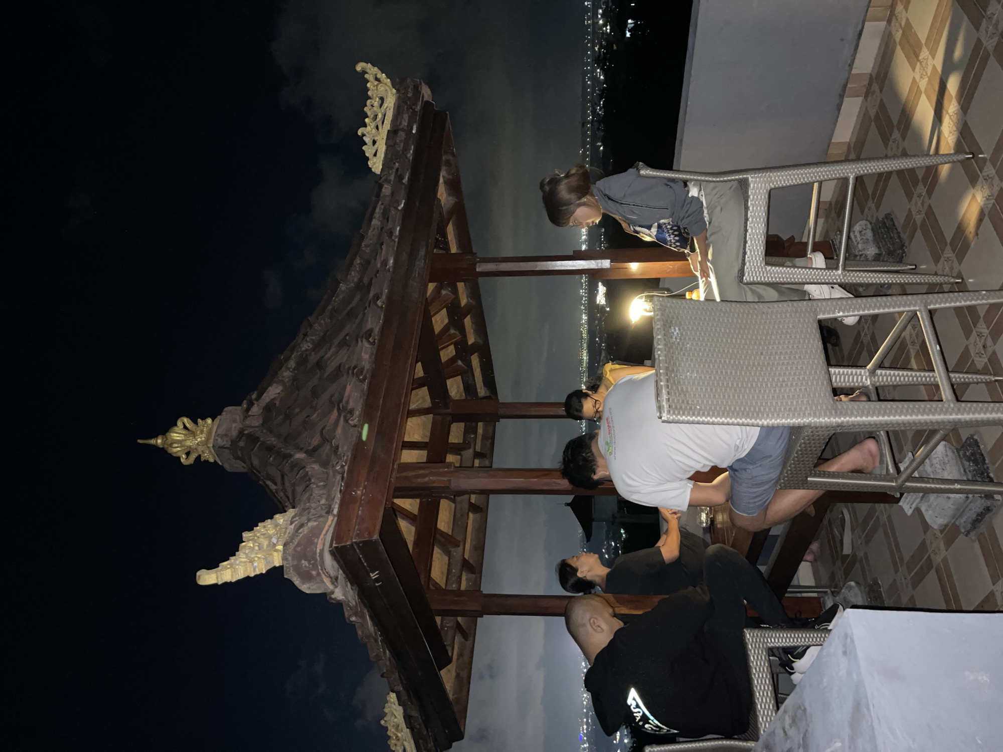 Four individuals are seated around a table under a traditional Balinese-style gazebo at night, overlooking a brightly lit cityscape which is Kuta Selatan, Indonesia. A man in a black hoodie is on the left, facing away from the camera. Next to him, a woman in a dark shirt is looking down. In the center, a man in a white t-shirt is also looking down. On the right, a woman in a grey jacket is looking to her left. The gazebo has a wooden structure with a tiled roof adorned with golden decorative elements. The surrounding area appears to be a rooftop or balcony with a railing. The lighting is dim, with a lamp on the table casting a yellow glow. The background cityscape is extensive with numerous lights.