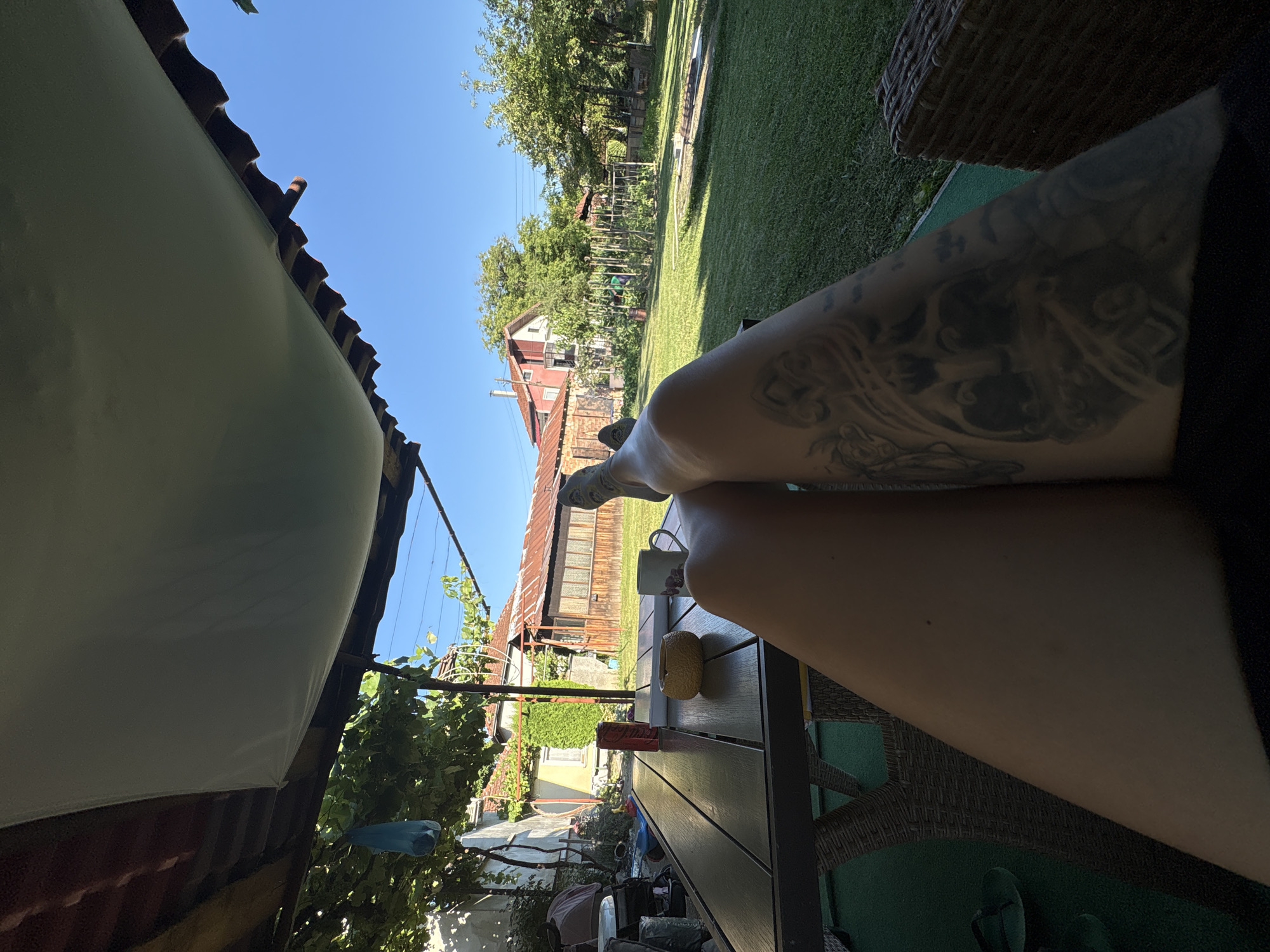 A first-person perspective shot captures a person's legs resting on what appears to be a woven chair, with a tattoo visible on the right leg. The scene is set outdoors, under a canopy, with a table in the foreground holding a can of Coca-Cola, an ashtray, and a mug. Beyond the table is a grassy backyard with trees and houses in the background. The sky is clear and blue. The image appears to be taken in Oreshak, Bulgaria, a village known for its pottery and woodworking traditions. The warm, sunny weather suggests it's a pleasant day. The overall atmosphere is relaxed and casual.