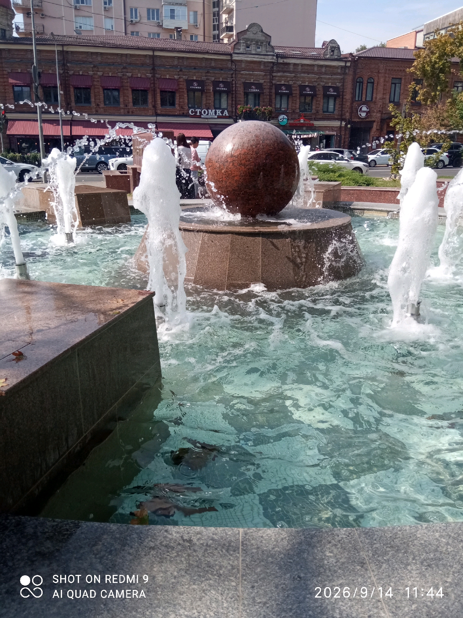 The image depicts a public fountain in the city of Rostov-on-Don, Russia, on a sunny day. The fountain features a large, reddish-brown spherical sculpture at its center, surrounded by multiple water jets that create a frothy, dynamic effect. The water is a clear, turquoise color, reflecting the sky and the surrounding architecture.
In the background, there are buildings with red brick facades, characteristic of historical architecture in the city. Some buildings have awnings and signs, suggesting they are commercial establishments, possibly cafes or shops. The sign "СТОМКА" is visible on one of the buildings. People, two women, are seen standing near the fountain, appearing to observe it or interact with each other. Several cars are parked or driving on the street behind the fountain, indicating an urban setting. The foreground of the image shows the edge of the fountain basin, made of granite-like material, with some fallen leaves on its surface. The overall scene suggests a pleasant, active public space in Rostov-on-Don.