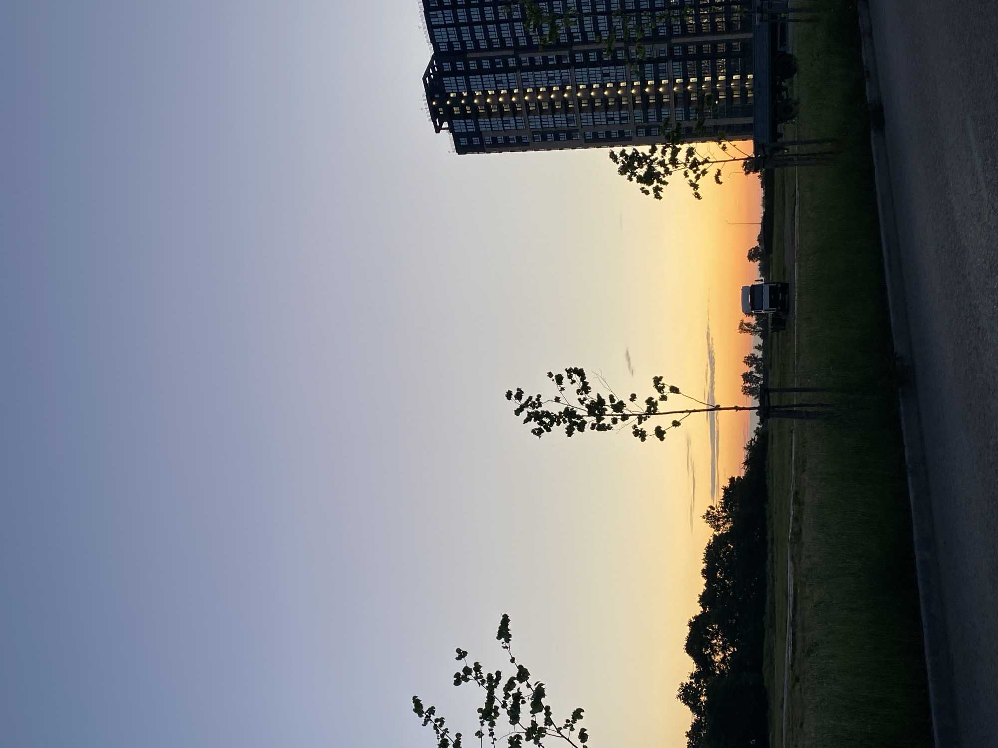 A high-angle shot captures an apartment building at dusk in Unknown, Unknown. The building, a tall, dark structure with numerous lit windows, dominates the right side of the frame. In the distance, silhouetted trees form a line across the horizon, with a truck and a flatbed trailer parked in a grassy field. The sky transitions from a clear, pale blue at the top to shades of orange and yellow near the horizon, indicating the sun has recently set. In the foreground, a patch of grass and a paved area lead the eye towards the scene. Two small trees with sparse leaves stand in the middle ground, one on the right and one on the left of the frame, with a third larger tree in silhouette on the far left.