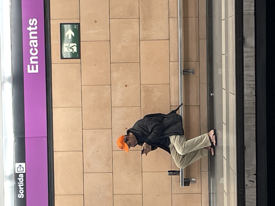 An adult male is seated on a metal bench on a subway platform in the Encants station in Barcelona, Spain. He wears an orange baseball cap, a dark jacket, light-colored trousers, and black flip-flops. A backpack is visible over his shoulders. He is actively engaged with a smartphone held in both hands. To his left, a black beverage can rests on the bench. The background consists of a tiled wall made of large, light brown rectangular tiles. Above the wall, a magenta sign displays "Encants" in white text and "Sortida" (Exit) with an arrow. A green exit sign with a running figure and an arrow is mounted on the tiled wall to the right. The platform floor is tiled, with a darker edge indicating proximity to the tracks. The track area below the platform is dark.