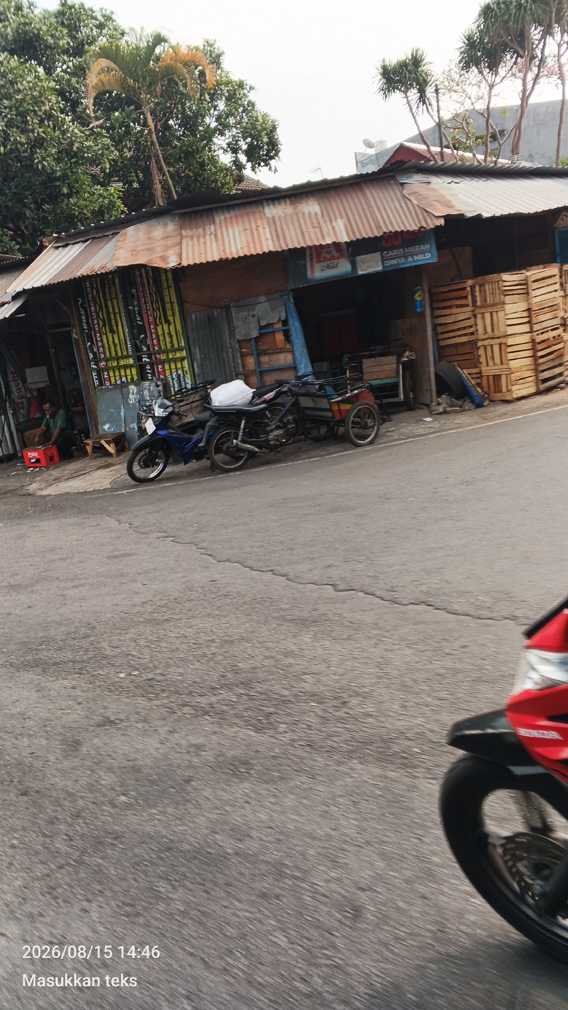 The image depicts a street scene in Malang, Indonesia, with a focus on two motorcycles parked in front of a shop. The shop has a corrugated metal roof and appears to be selling various goods, with stacks of wooden crates visible to the right. A person is seated near the entrance of the shop, beside a red crate. One motorcycle is blue and white, while the other is black and has a sidecar. The road in the foreground is asphalt, and the image has a motion blur, suggesting it was taken from a moving vehicle. The timestamp "2026/08/15 14:46" and the text "Masukkan teks" (Insert text) are overlaid on the bottom left of the image. Trees and buildings are visible in the background, indicating an urban or semi-urban environment.