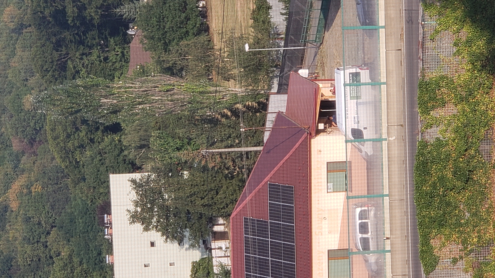 The image shows a suburban scene in Prešov, Slovakia, dominated by a residential building and a smaller commercial structure. The residential building, a modern, white, multi-story structure with visible windows, is partially obscured by trees. To the right, a single-story commercial building with a reddish-brown roof is prominent. This building features a section with solar panels installed on the roof. In front of the commercial building, there is a road or parking area with a concrete barrier and a green fence. Two vehicles are visible: a silver van and a white panel van. The white van has lettering on its side, indicating a business, possibly related to electrical services or installations. Two individuals are partially visible near the entrance of the commercial building, one standing and the other seated. The background consists of a densely wooded hillside. The overall atmosphere suggests a sunny day in a developed area adjacent to a natural landscape.