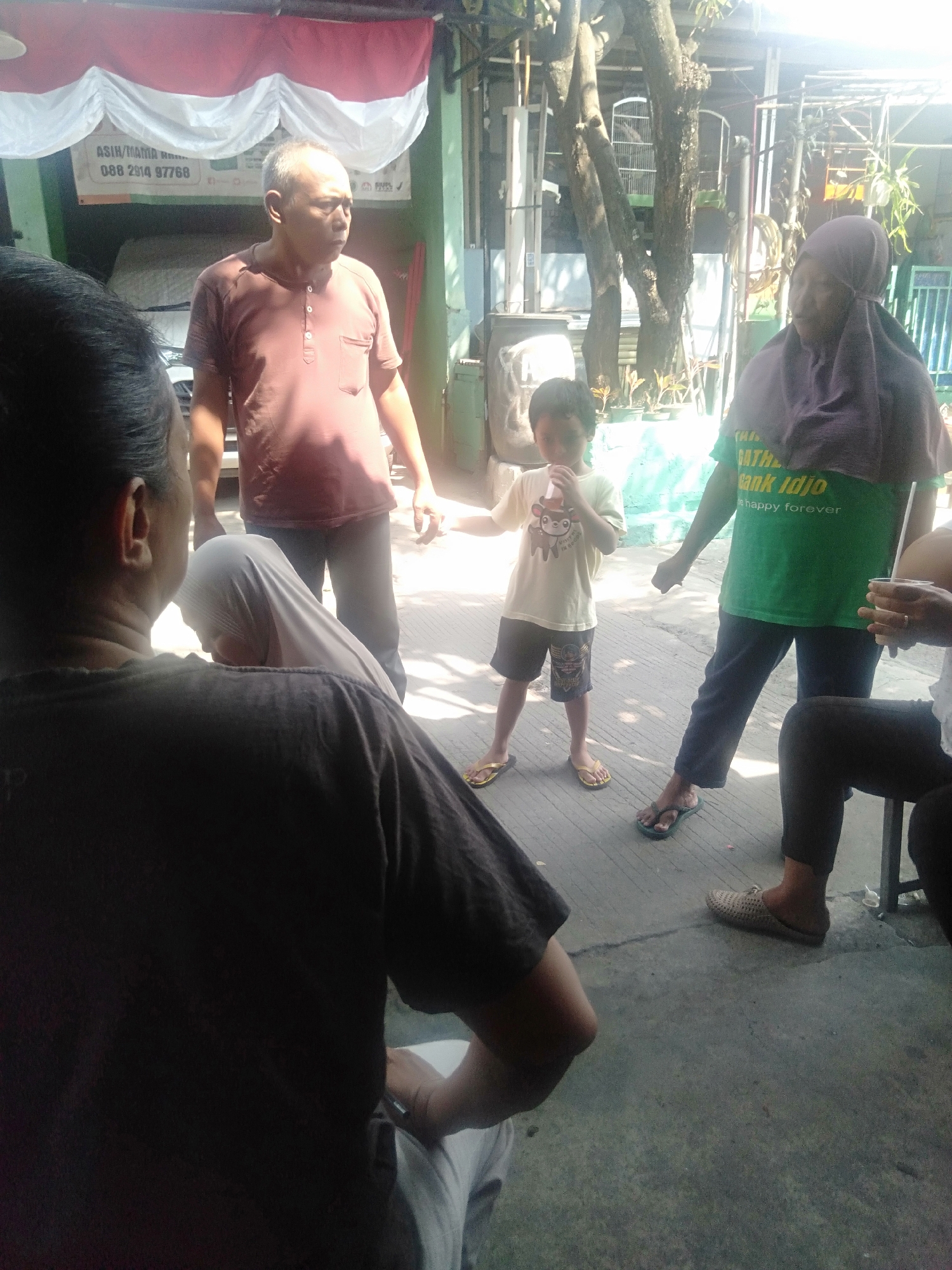 In Ciantra, Indonesia, an outdoor gathering occurs under a bright, sunny sky, with a large white and red banner prominently displayed overhead. The scene features a group of people, including a young boy in a light-colored t-shirt and dark shorts, holding a drink with a straw. Beside him, a woman wears a green t-shirt with text and a purple headscarf. Further to the left, a man in a reddish-brown t-shirt faces away from the camera. In the foreground, a person wearing a dark t-shirt sits, with their back to the viewer, and another individual, partially visible in a light-colored garment, is seated to their left. The ground is a concrete surface, and trees with visible branches are present in the background, suggesting an open, possibly semi-public, space.
