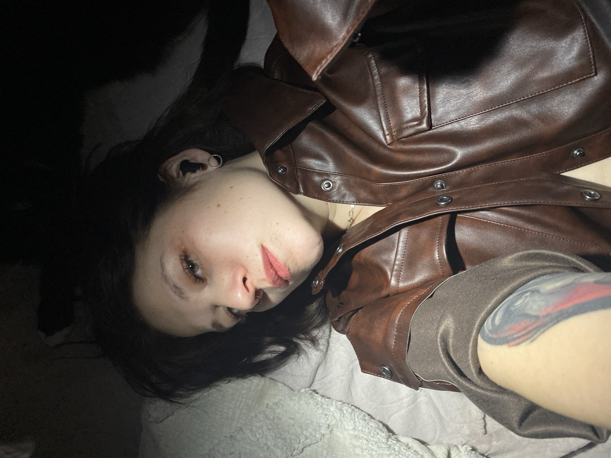 This media file presents an angled, close-up portrait of an individual lying down indoors. The subject, a young person with dark hair, fair skin, and freckles, gazes directly towards the camera with a relaxed or pensive expression. They are dressed in a brown, leather-look jacket with visible snap fasteners and a brown fabric lining on the sleeve. Notable accessories include a silver hoop earring and a dark stud in the ear, along with a nose ring. A colorful tattoo, featuring red, black, and grey tones, is partially visible on the person's left arm.
The individual is positioned on a light-colored, textured surface, possibly a blanket or sheet, suggesting an indoor setting like a bedroom or living space. A very dark, indistinct shape is present in the upper left background, which might be a pet.
The lighting is dramatic, with strong highlights on the subject's face and jacket contrasting with deep shadows, indicating a dim environment, possibly evening or nighttime, with an artificial light source. The image is composed at an angle, with the subject's head towards the bottom left and body extending to the upper right. There is no visible text in the image. The context provided for this media file is Chaikovsky, Russia.