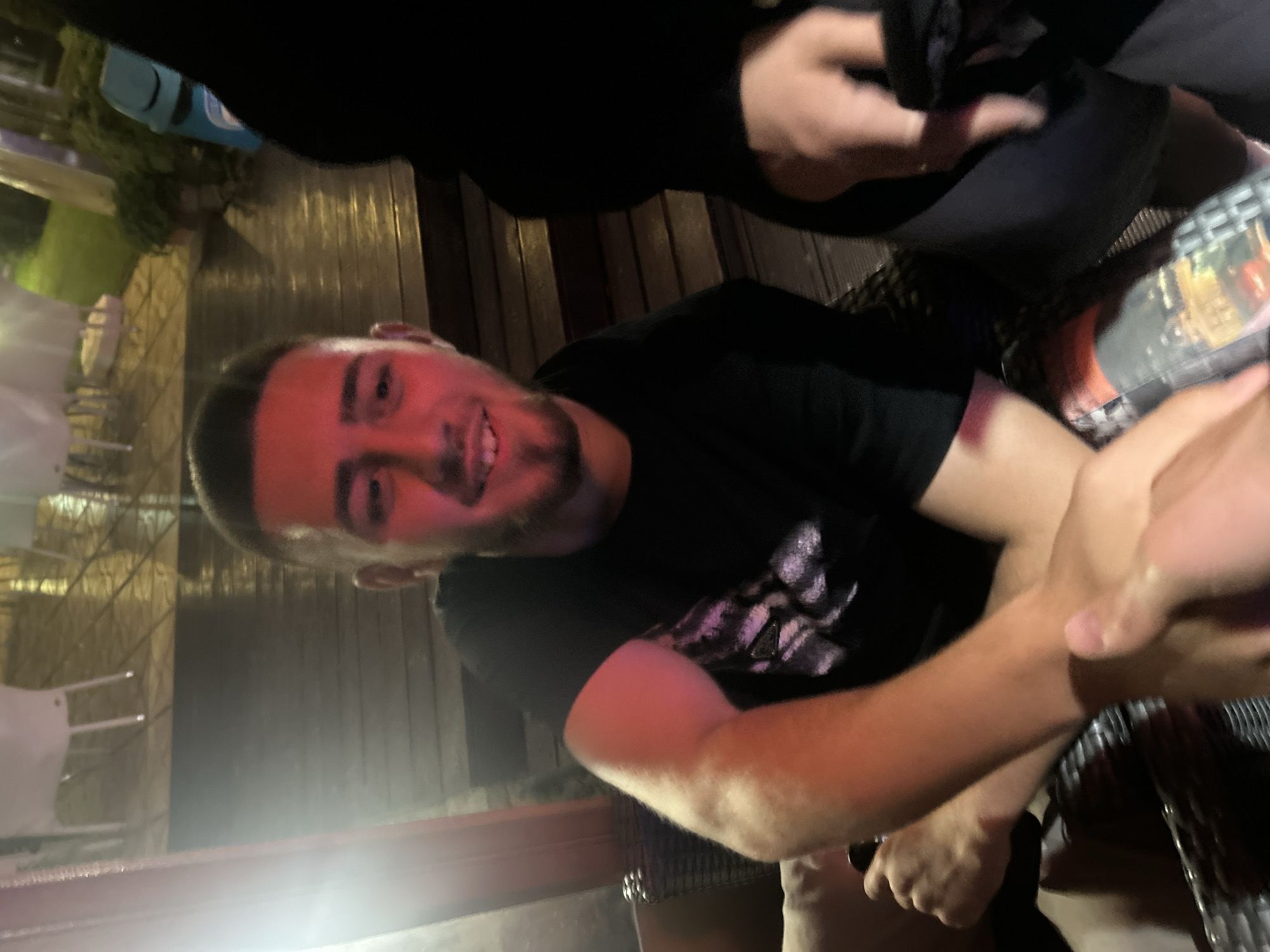 A man with short brown hair and a beard is at an outdoor table, smiling at the camera. He is wearing a black t-shirt. Another person is next to him, with a hand near the table. The table appears to be wooden, with a reflective surface. There is a building and greenery in the background. A blue object can be seen on a nearby table. The location is likely a restaurant or bar in Elhovo, Bulgaria.