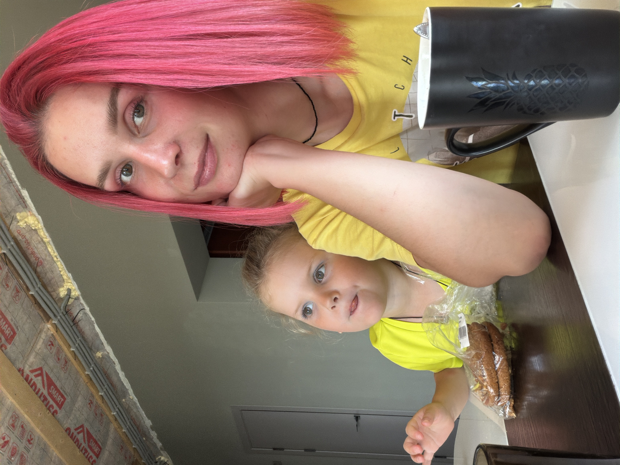 The image shows two people, an adult with bright pink hair and a young child, in what appears to be an indoor setting. The adult is resting her chin on her hand, looking towards the camera with a slight smile. She is wearing a yellow t-shirt. Beside her, there is a black mug with a pineapple design. The child, also wearing a yellow t-shirt, is looking at the camera with a curious expression and has their tongue sticking out slightly. In front of the child, there is a plastic bag containing what looks like bread rolls or pastries. The background shows a wall, part of a ceiling, and some construction materials with visible insulation and electrical wiring. The lighting suggests it is daytime. The overall mood appears relaxed and casual, possibly a moment captured during a break or snack time. There is some visible text on the construction materials, including "SMART" and "AUDITISS." The location context is Chaikovsky, Russia.