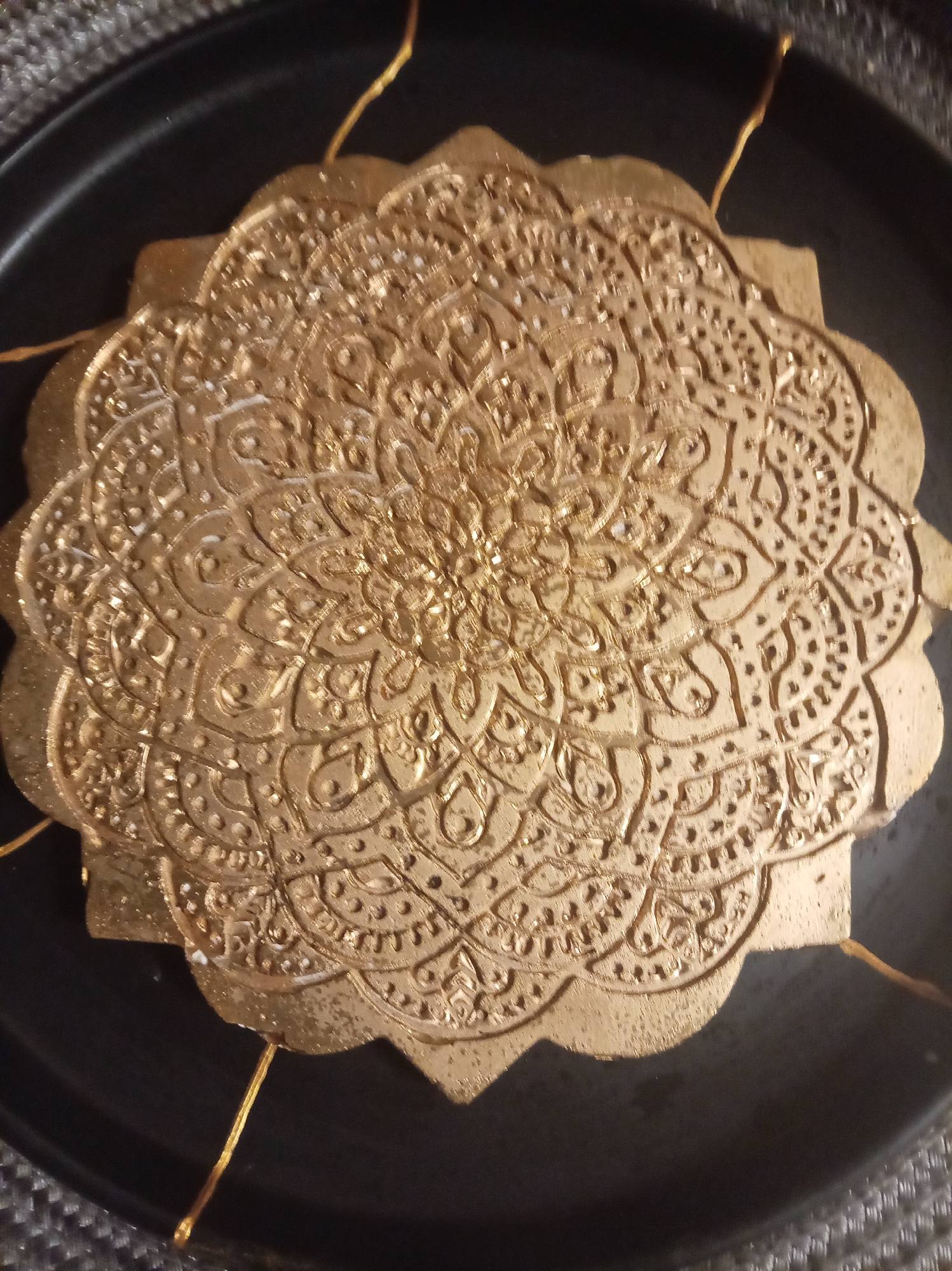 A close-up, overhead shot shows a golden, circular, embossed decorative item placed on a dark, round surface. The decorative item has intricate, symmetrical patterns resembling a mandala or a lace doily. The edges of the decorative item are scalloped. Several thin, golden threads are attached to the item and extend outwards, some disappearing beyond the frame. The dark surface appears to be a plate or tray with a textured rim. The overall impression is of an artistic or craft object, possibly a cookie or a piece of art. The location in Yessentuki, Russia is not determinable from the image.