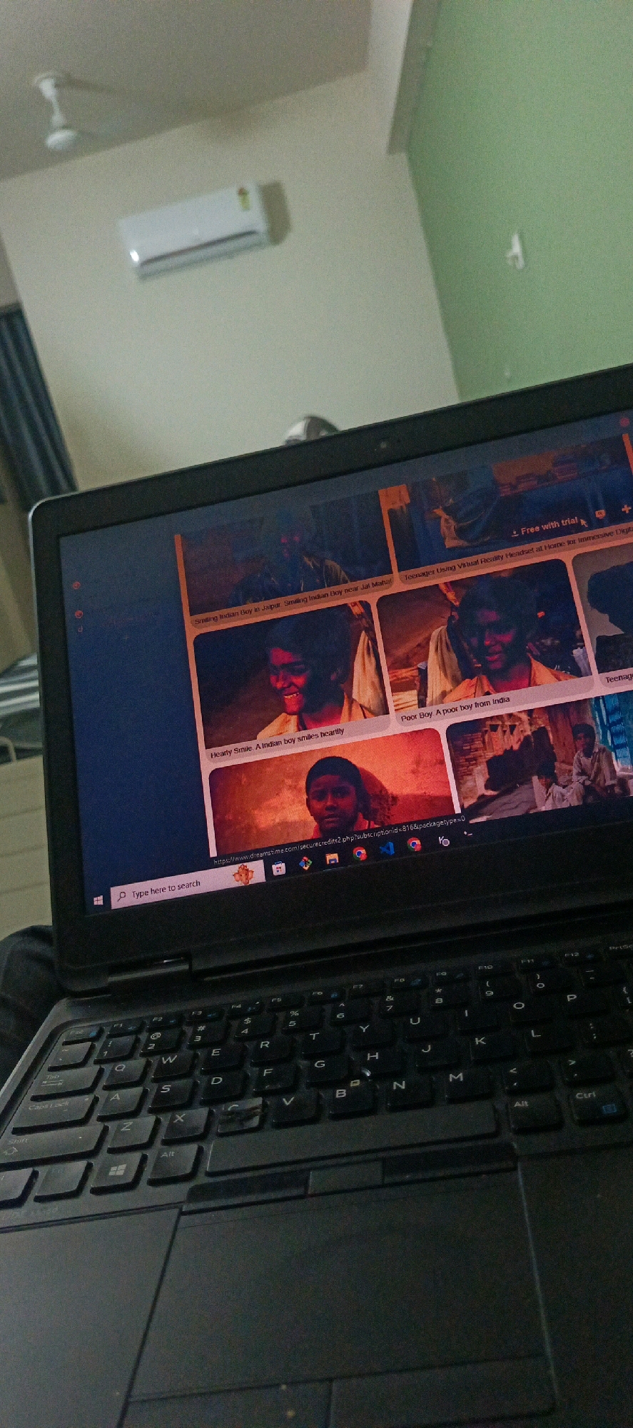 The image shows a laptop open on a dark surface, likely a table or desk. The laptop screen displays a webpage with multiple images of young Indian boys. The webpage appears to be a stock photo website, with captions like "Smiling Indian Boy in Jaipur," "Hearty Smile. A Indian boy smiles heartily," and "Poor Boy. A poor boy from India." The URL visible on the screen suggests it's from dreams time.com. The environment outside the laptop screen is dimly lit and appears to be an indoor setting. A white ceiling fan is visible on the ceiling in the upper left corner, along with a white air conditioning unit mounted on a light-colored wall. A green wall is visible in the background on the right. The keyboard of the laptop is clearly visible in the foreground. The image is taken from a slightly elevated angle, looking down at the laptop. The lighting suggests it may be evening or a room with limited natural light.