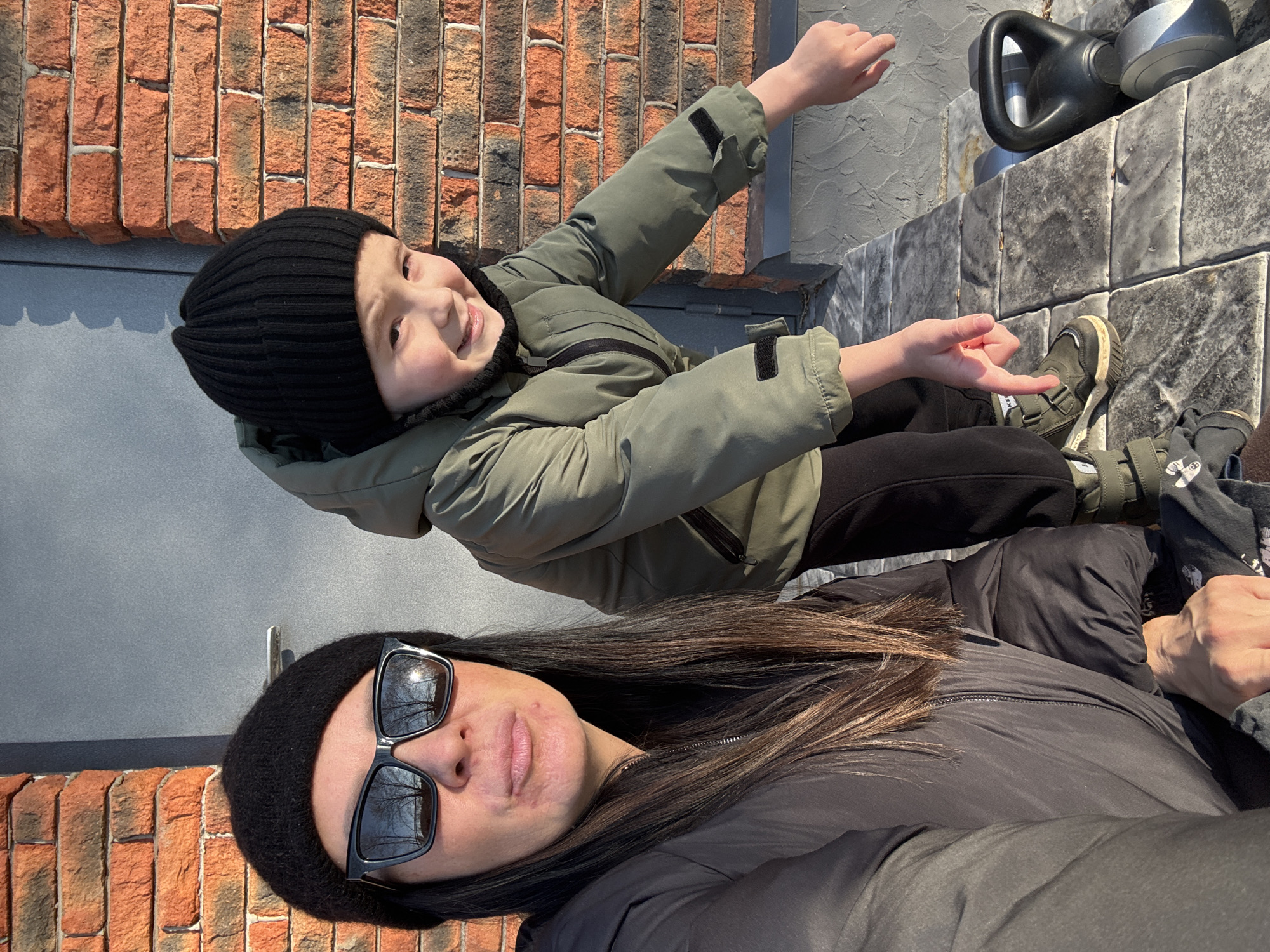 A selfie captures a woman with long brown hair wearing sunglasses and a black beanie, alongside a child in a green jacket and black beanie. The child is positioned above and behind the woman, reaching an arm out towards a grey watering can situated on grey tiled steps. The background features a brick wall on the left and a grey wall to the front of the left of the image. The location is Chaikovsky, Russia.