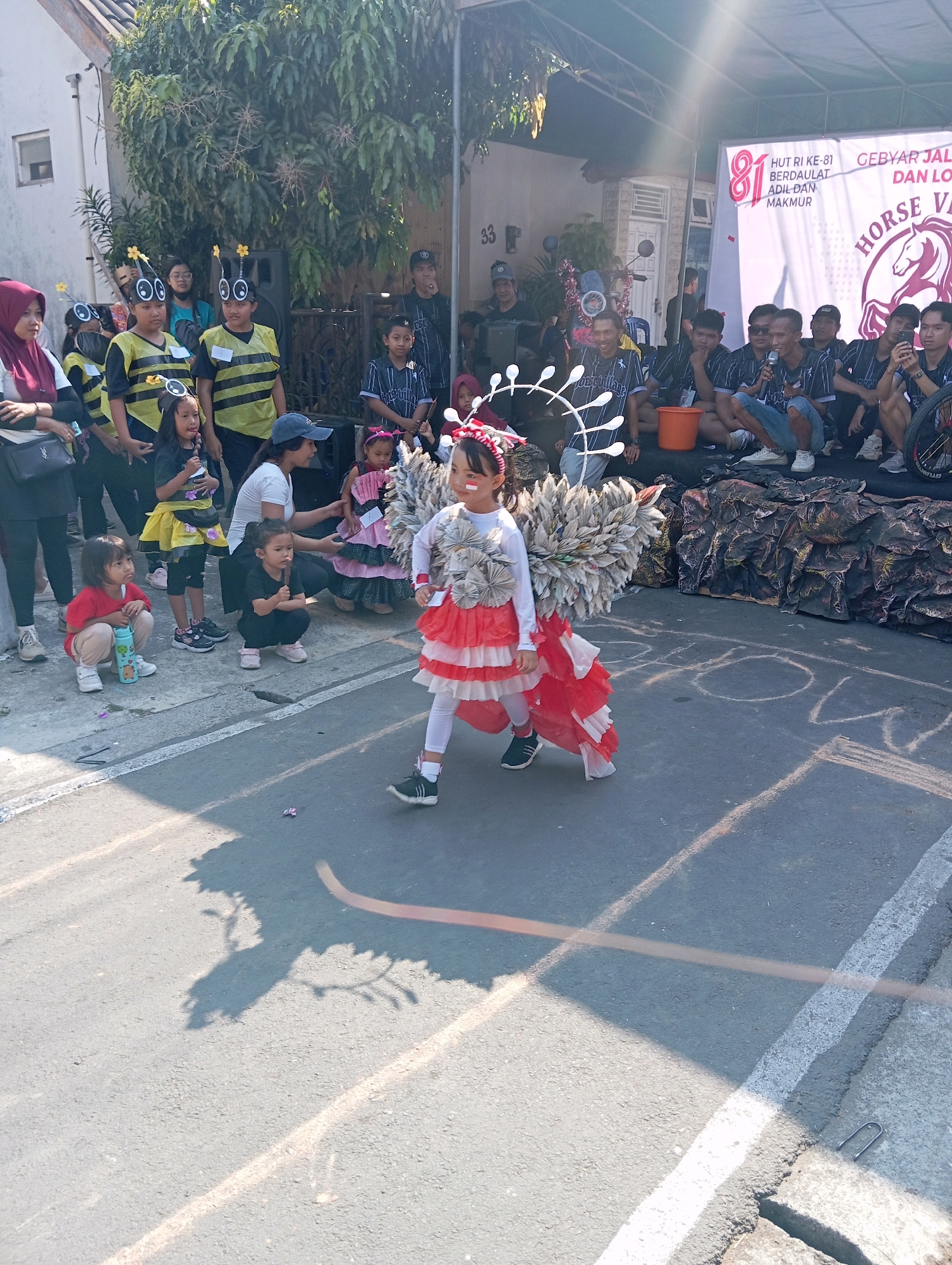 The image depicts an outdoor street event in Magelang, Indonesia, likely a parade or festival. In the foreground, a young girl in a costume with white and red ruffles and a large, feathery headdress walks down the street. She has red and white face paint. Several other children are seated or standing nearby, some wearing bee-themed costumes. Adults are visible in the background, including a group sitting on a stage behind a banner. The banner has text in Indonesian, including "HUT RI KE-81" (81st anniversary of the Republic of Indonesia) and "GEBYAR JALAN" (Street Festival). The scene is illuminated by bright sunlight, casting shadows on the asphalt road. Buildings and trees form the backdrop.