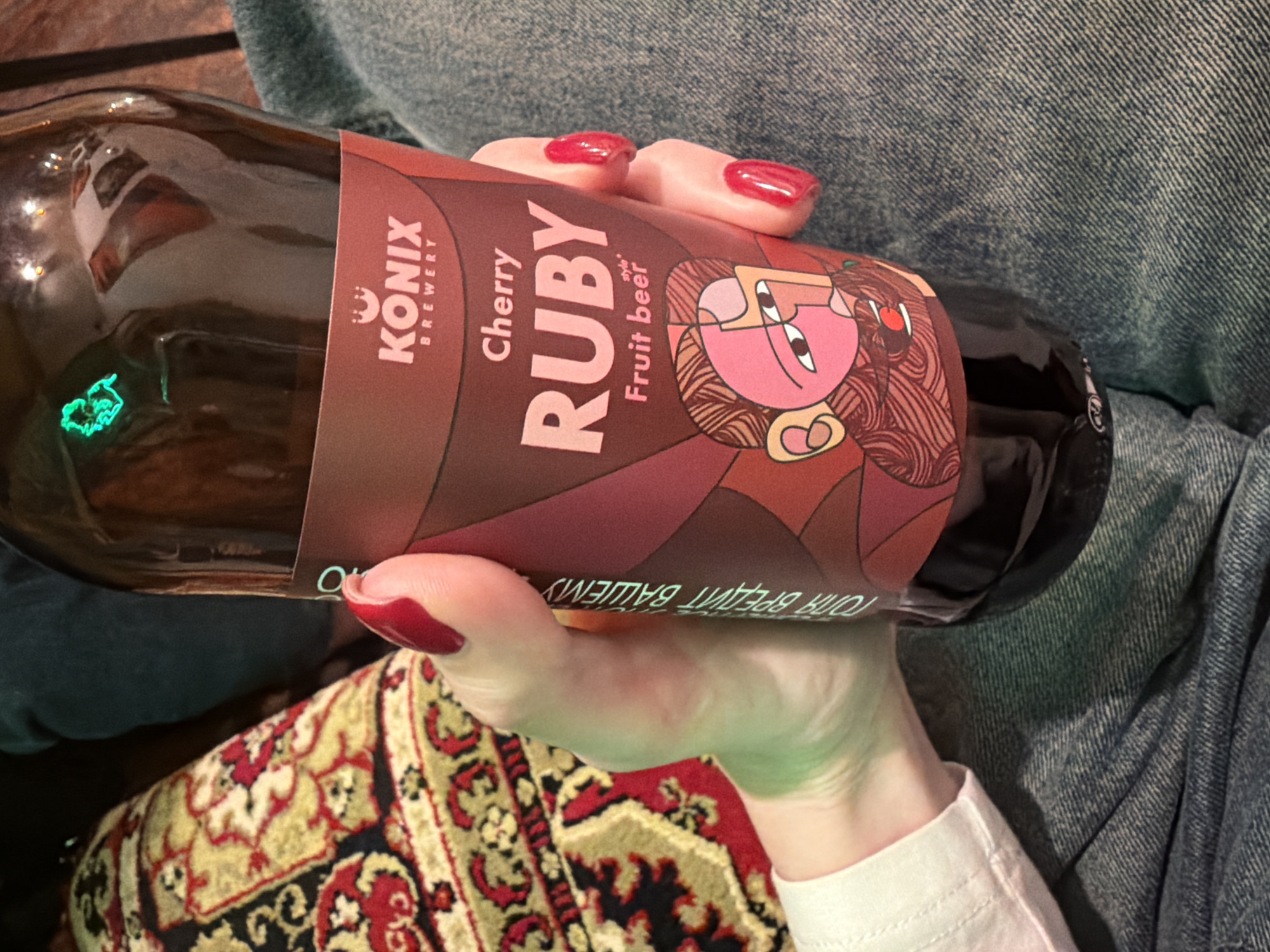 A person is holding a bottle of Konix Brewery Cherry Ruby Fruit Beer. The label features the Konix Brewery logo and an illustration of a stylized face. The person has red nail polish. The scene includes a portion of a colorful patterned rug and the person's jeans. The location is Rostov-on-Don, Russia.