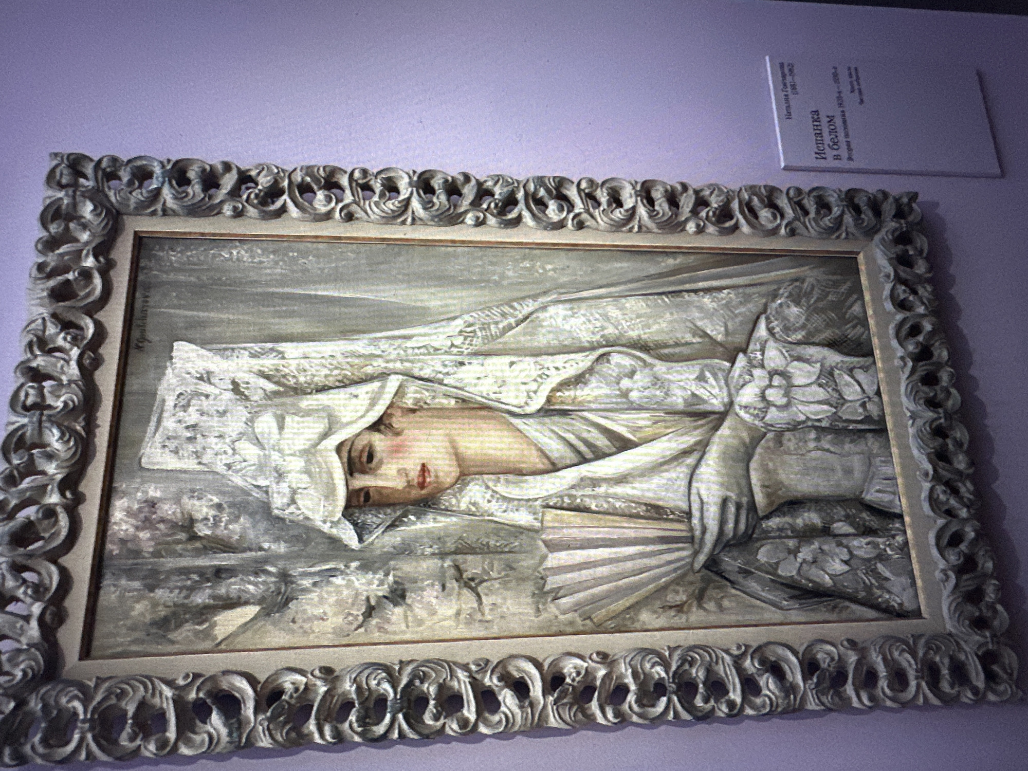 An oil painting titled "Spain in White" by Natalya Goncharova, dating from 1930-1932, is displayed in a decorative, ornate frame. The painting depicts a woman in a white, lace-like headdress and a white dress, holding an open fan. The background is rendered in muted tones of gray and purple, with abstract shapes that suggest fabric or drapes. The overall style of the painting is modern, with bold brushstrokes and a somewhat flattened perspective. A white informational plaque is attached to the wall to the right of the painting, providing details about the artwork and artist, including "Наталья Гончарова (1881-1962)", "Испанка в белом", and "1930-1932". The painting is hung on a lilac-colored wall. This artwork is located at the Balashikha Museum of History and Local Lore in Balashikha, Russia.