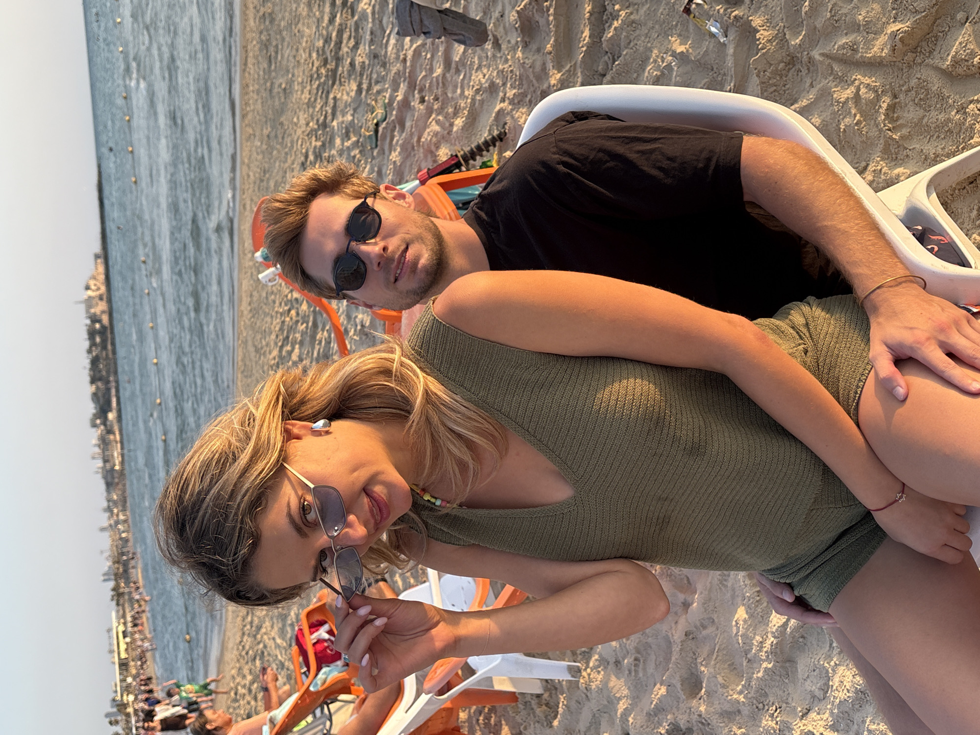 A woman sits on the lap of a man on a sandy beach during the golden hour in Tel-Aviv, Israel. The woman, with blonde hair, wears a V-neck olive green knitted top and matching shorts. She holds silver-framed sunglasses, looking over them at the camera. She also wears silver hoop earrings, a colorful beaded necklace, a thin bracelet on her right wrist, and a red string bracelet with a Star of David charm on her left wrist. The man, with light brown hair and stubble, wears dark sunglasses and a black t-shirt, seated in a white plastic chair. His left hand rests on the woman's left thigh, and he looks at the camera. In the background, other individuals are visible on the beach and in the water, alongside orange and white plastic beach chairs. The Mediterranean Sea has visible buoys marking an area. Beyond the water, a cityscape consistent with Jaffa, part of Tel-Aviv, is discernible under a clear sky.