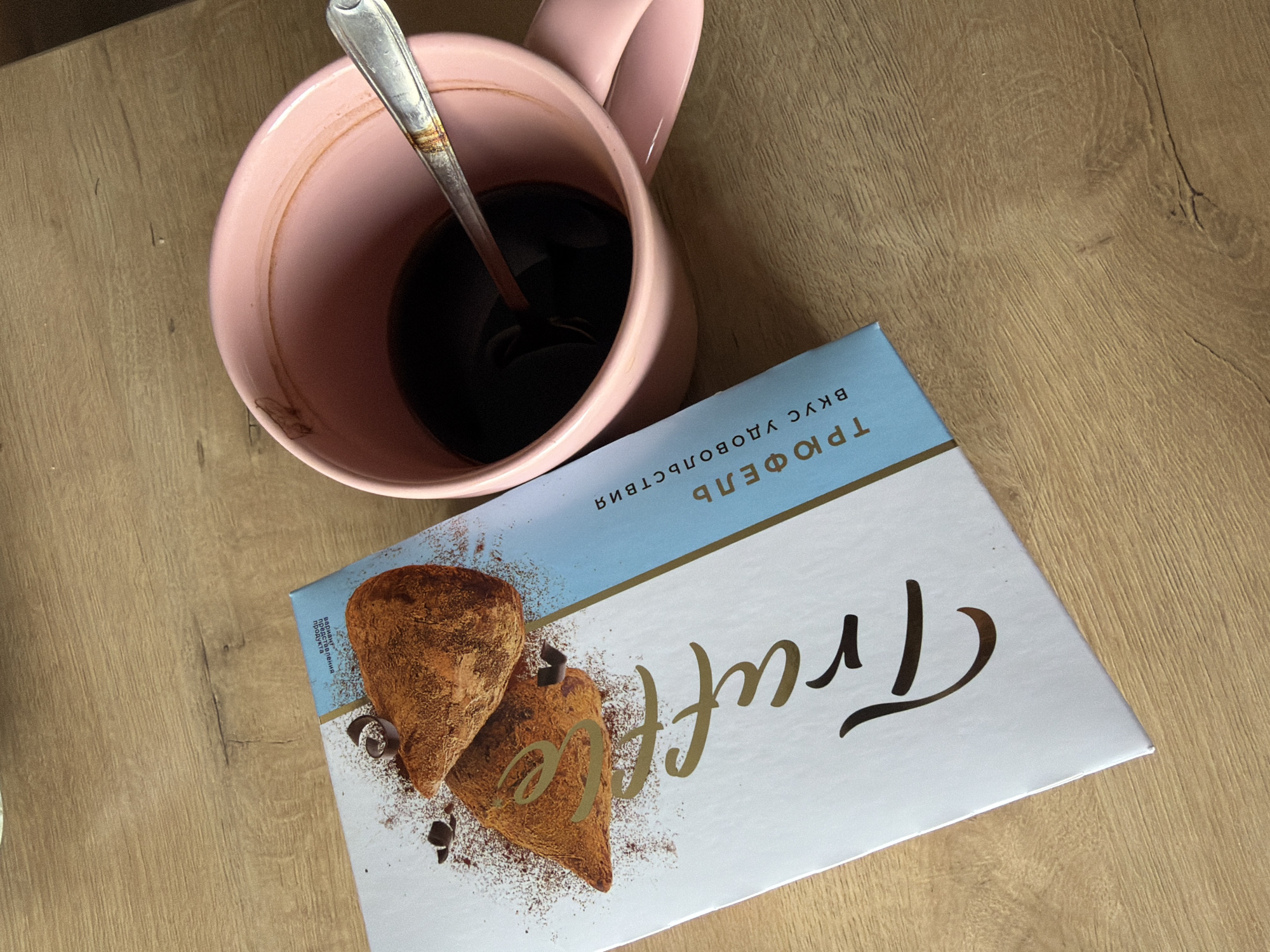 This image shows a close-up overhead view of a wooden surface. On the left is a light blue and white box with text in gold and black, and an image of two chocolate truffles. The text on the box appears to be in Russian and includes the word "Truffle". To the right of the box is a pink mug filled with a dark liquid, presumably coffee, with a silver spoon resting inside. The mug has some residue on the inside rim. The scene is lit by natural light, casting shadows on the wooden surface. The image does not contain any elements that would indicate its location in Ust-Ilimsk, Russia.