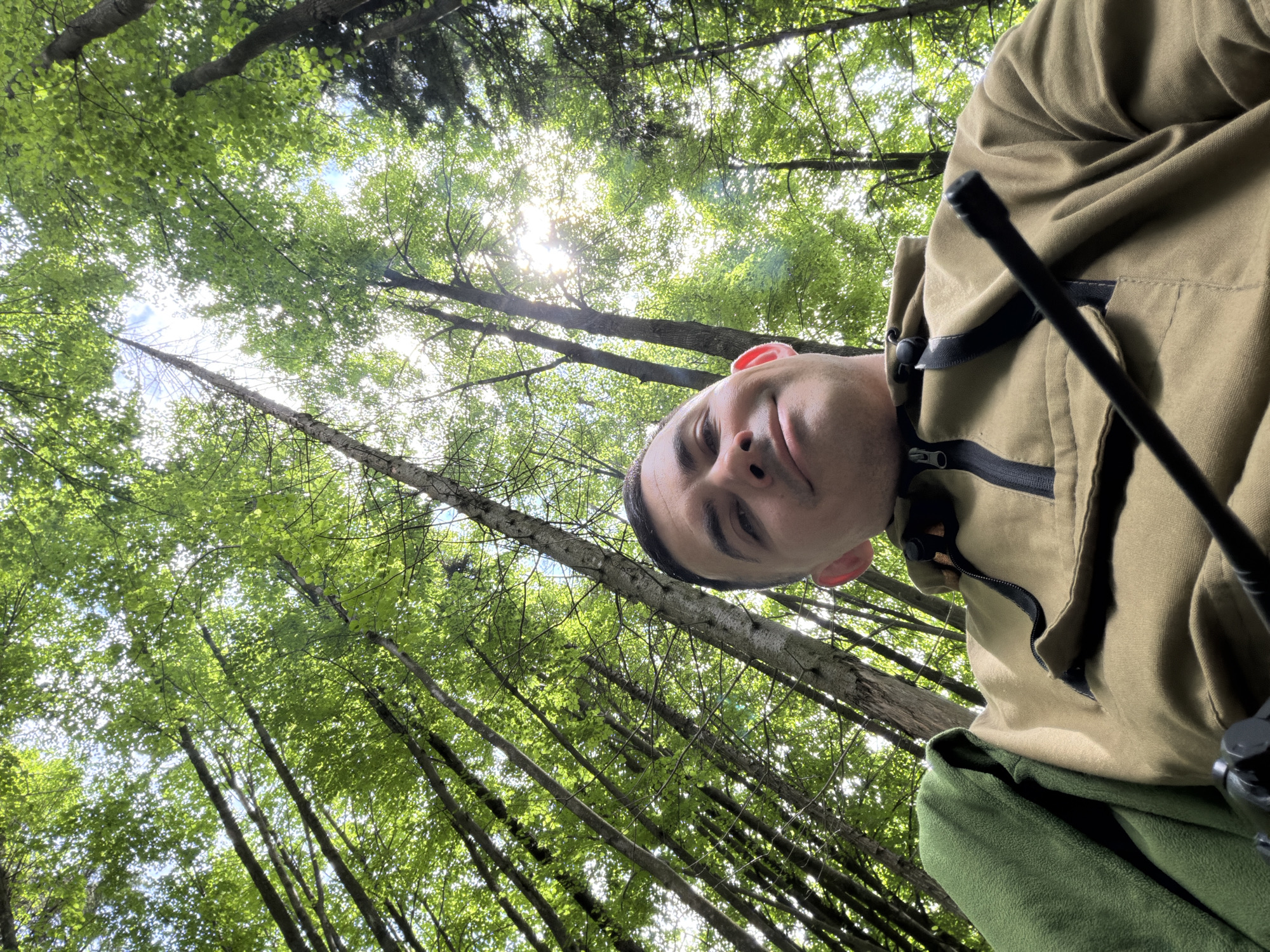 A male individual is depicted in a low-angle photograph, framed against a backdrop of dense forest foliage under bright daylight in Kechur, Russia. The man, with short dark hair and fair skin, appears to be in his 20s or 30s, looking upwards and exhibiting a slight smile. He is wearing a light khaki or brown anorak-style pullover jacket featuring a black chest zipper and a horizontal black stripe. A green fleece garment is draped over his left shoulder. In the lower right foreground, a black, slender, cylindrical object, possibly a stick or part of a device, extends diagonally. The environment consists of numerous tall, slender tree trunks with vibrant green deciduous leaves forming a dense canopy overhead. Bright sunlight penetrates the foliage, creating illuminated patches and visible lens flare, with glimpses of a clear blue sky through the gaps. The perspective is upward-looking, capturing the individual and the forest canopy.