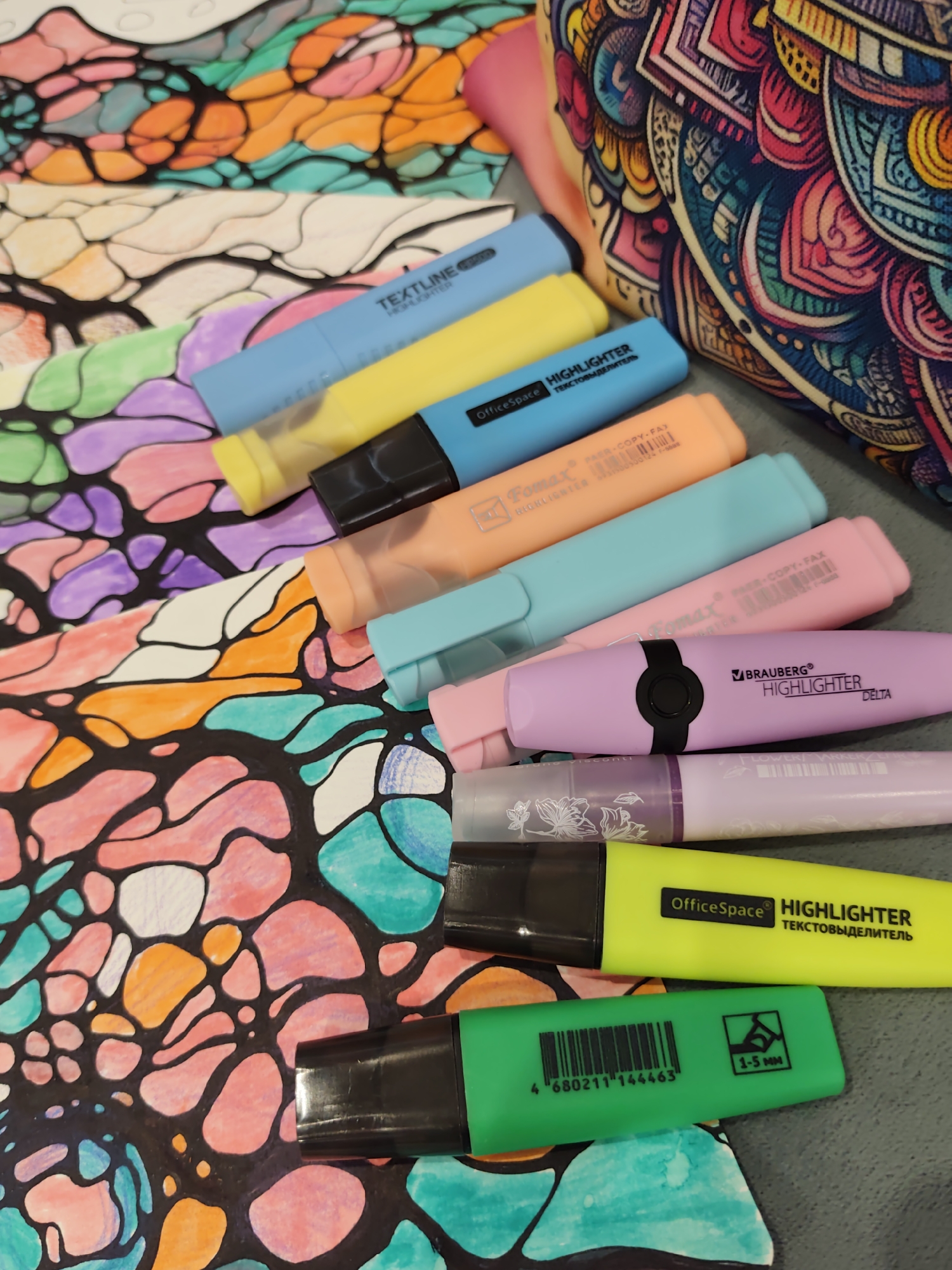 The image depicts a collection of highlighters laid out on a surface, with a colorful abstract drawing underneath. The highlighters are of various colors, including blue, yellow, orange, light blue, pink, purple, light purple, and green. Some of the highlighters have visible branding such as "TEXTLINE," "OfficeSpace," "Fomax," and "BRAUBERG." One of the highlighters has a label indicating "1-5 MM." In the background, a portion of a bag or fabric with an intricate, mandala-like pattern in vibrant colors is visible. The drawing beneath the highlighters features organic, interconnected shapes in shades of pink, orange, green, and teal, outlined in black. The surface appears to be a flat, grey surface, possibly a table. There are no people or animals present in the image. The scene is static and focuses on the arrangement of art supplies.
The image does not contain any information that would allow for its location to be determined, including in the city of Sochi, Russia.