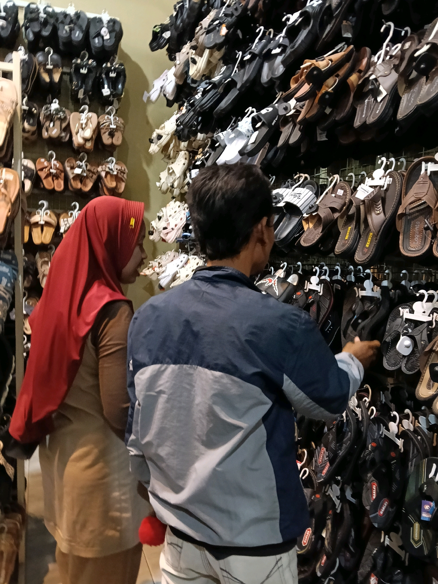 In a shoe store in Dengok, Indonesia, a woman wearing a red hijab and a tan long-sleeved shirt stands beside a man wearing a blue and gray jacket and white pants. They are positioned in front of a wall display filled with various types of sandals and flip-flops. The sandals are predominantly dark-colored, with some lighter brown and tan options also visible. The display consists of multiple rows of footwear, with each pair hanging from a hook. The lighting in the store appears to be artificial and somewhat dim. The overall impression is a typical retail environment focused on footwear sales.