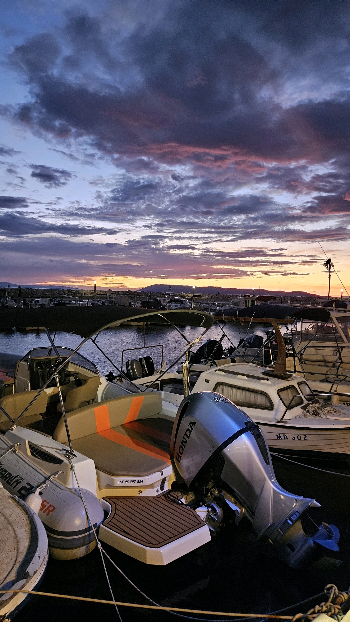 The image depicts a marina at sunset in Općina Tučepi, Croatia. The sky is filled with dramatic clouds displaying hues of purple, pink, and blue, illuminated by the fading sunlight. Several boats are docked in the foreground, with their engines visible. The closest boat features a silver Honda outboard motor, a tan and orange striped seat, and a textured brown deck step. Another boat behind it has "MA 302" visible on its hull. In the background, a line of boats stretches across the water, with a breakwater and distant hills forming the horizon. A single palm tree stands silhouetted against the colorful sky, and a lighthouse or navigational marker is visible near the breakwater. The water is calm, reflecting the sky's colors. The scene is quiet and serene, suggesting the end of a day's activities in the marina.