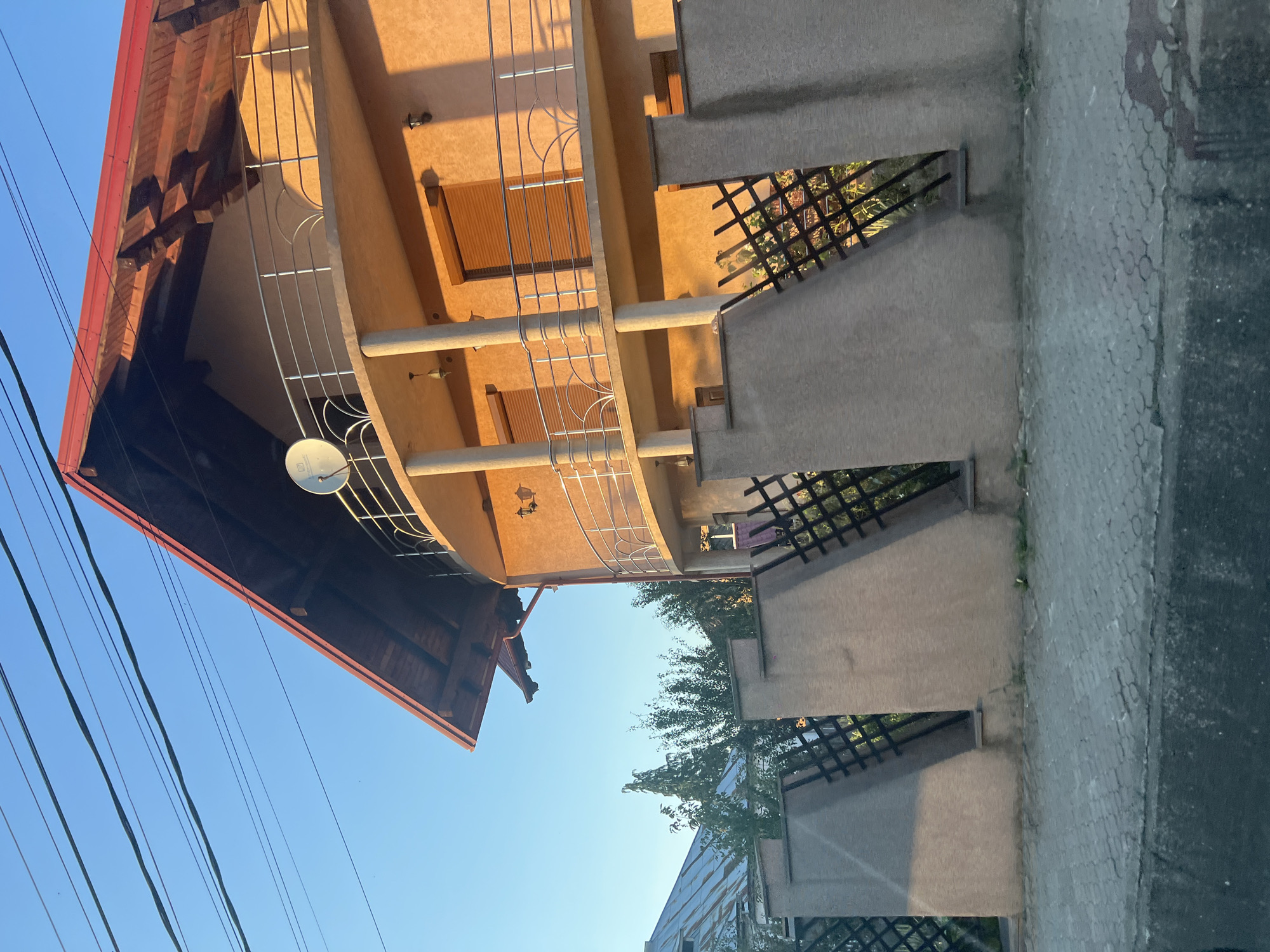 The image depicts a segment of a two-story residential building with an orange stucco exterior, situated in Rona de Sus, Romania. The building features a red tile roof, a rounded second-story balcony with silver railings, and multiple windows with brown rolling shutters. A satellite dish is mounted on the upper portion of the building. A concrete fence with integrated decorative black lattice work separates the property from the street. The scene is illuminated by the warm light of either sunrise or sunset, casting long shadows across the fence and pavement. Overhead, a tangle of power lines is visible against a clear blue sky. The foreground shows a section of a paved sidewalk and what appears to be a street or road. Trees are visible behind the fence, and another building with a metal roof is partially visible to the left. There are no people or animals visible in the frame.