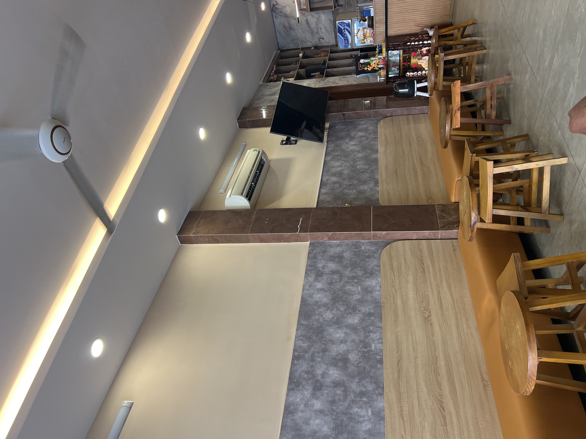 The image captures the interior of a restaurant or bar in Nha Trang, Vietnam. The scene is dominated by a layout featuring tables and chairs. Wooden stools surround a long table. On the left side of the image is a beige wall with a dark brown accent border, upon which a flat-screen TV is mounted. An air conditioning unit is present near the TV.
The floor consists of gray tile. The ceiling is white, featuring recessed lighting and a ceiling fan. In the background, shelves and cabinets are seen with what appears to be various beverages displayed, suggesting a bar area.