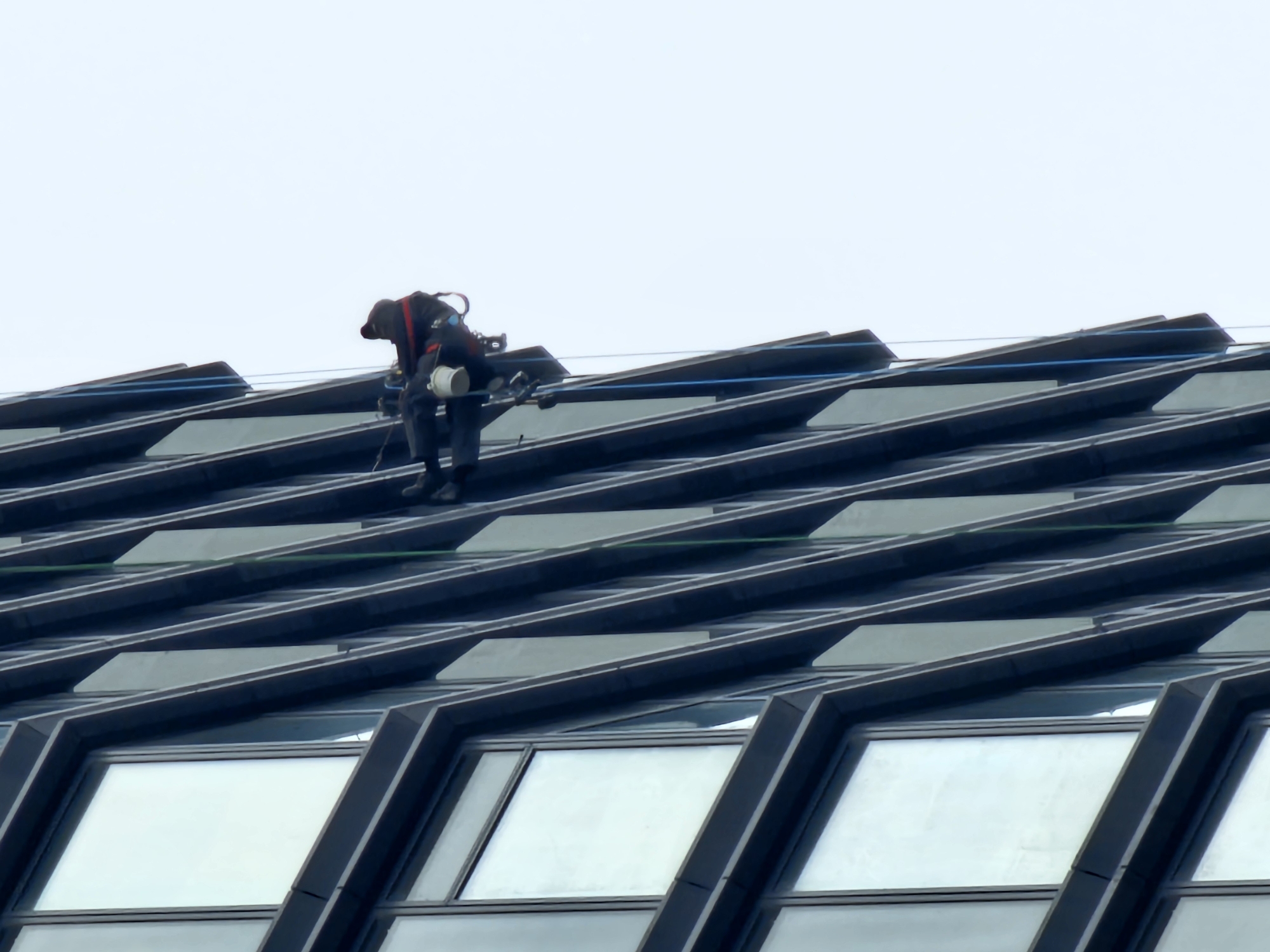This media file depicts a worker on the steeply angled roof or upper facade of a modern building in Moscow, Russia.
The primary subject is a person, likely a maintenance worker or window cleaner, positioned on the sharp incline of the building's structure. They are dressed in dark work clothing and appear to be wearing a safety harness, with a white cylindrical object (possibly a bucket or tool container) attached near their hip. Their posture suggests they are actively engaged in a task, bending forward over the surface.
The setting is the exterior of a contemporary architectural structure, characterized by its striking geometric design. The building's facade is composed of repetitive, angular sections of dark panels and glass windows, creating a pattern of parallel diagonal lines that extend across the frame. Blue and green safety lines or ropes are visible stretched across the facade, indicating the safety setup for the worker. The windows reflect the bright, overcast sky.
The activity taking place is the cleaning or maintenance of the building's exterior. The worker is performing this task at a significant height on a challenging, sloped surface, relying on safety equipment.
Notable details include the overcast sky, which provides soft, diffused lighting and suggests it is daytime. There are no strong shadows, indicating a cloudy or hazy day. The location is Moscow, Russia, as specified. No explicit emotions are discernible from the worker's distant figure. There is no visible text in the image. The image emphasizes the scale of modern architecture and the specialized work required to maintain it.