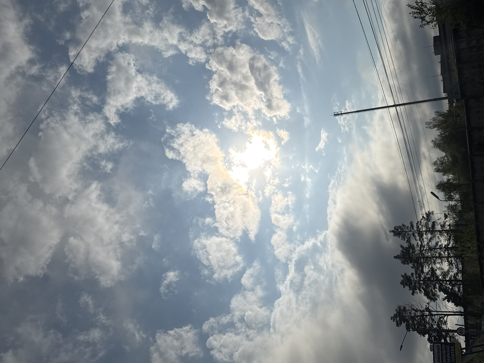 The image shows a sky with scattered white clouds and the sun partially obscured by the clouds. The sky is blue in the areas not covered by clouds. In the lower right corner, there are trees and some infrastructure, including poles and wires. In Russian, a sign can be seen. This image appears to be taken in the city of Chaikovsky, Russia.