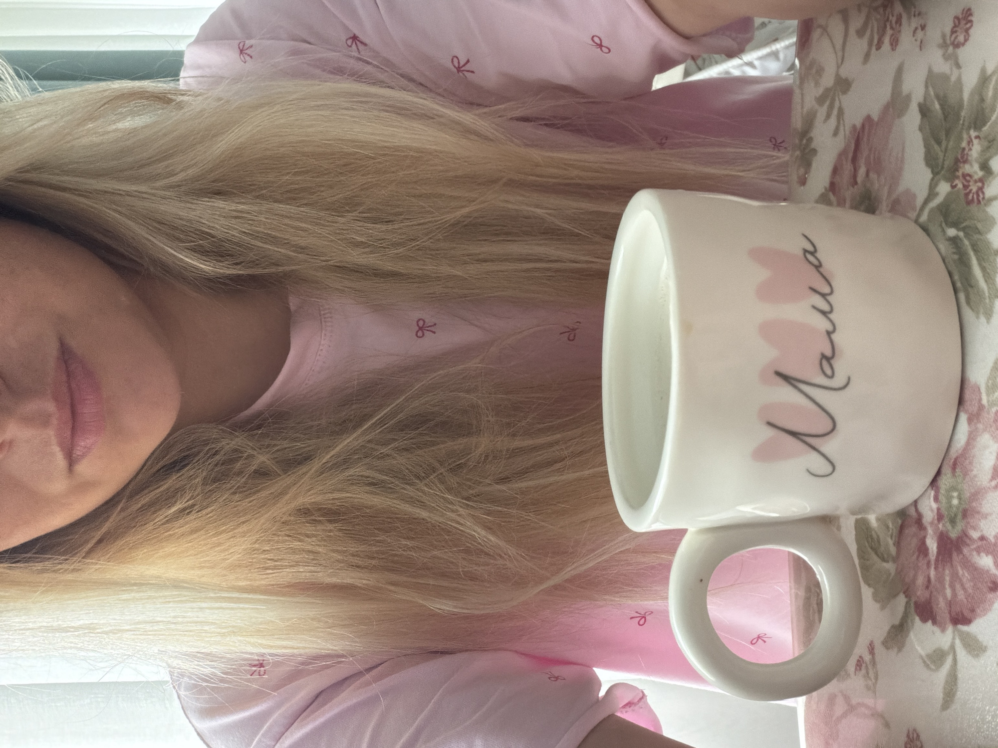 The image shows a close-up view of a person's lower face and upper torso, with their blonde hair falling forward. They are wearing a pink t-shirt adorned with small red bow patterns. In front of them is a white ceramic mug with a large, circular handle. The mug has the word "Mama" written on it in black script, accompanied by two pink heart shapes. The mug rests on a tray with a floral pattern featuring pink and green flowers. The scene appears to be indoors, possibly near a window with sheer curtains, suggesting a domestic setting. The provided metadata does not contain information about the specific location in Votkinsk, Russia.