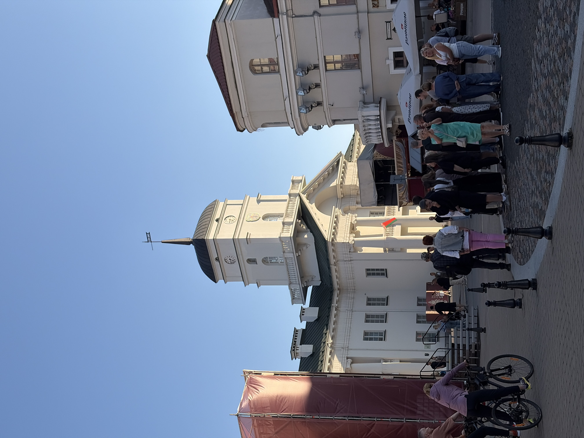 In this outdoor scene, a group of people are gathered in a public square in Minsk, Belarus, in front of a large white building with a clock tower. The architecture suggests a historic or administrative building. The sky is a clear, bright blue, indicating pleasant weather.
On the left side of the image, a person on a bicycle is visible, and further left, a large red structure, possibly part of a stage or scaffolding covered with fabric, is set up. A group of people are standing and interacting, some are looking at their phones, others are conversing. A woman in a green dress is holding up her phone, appearing to take a photograph. Several individuals are dressed casually, with a mix of men and women of various ages.
The ground is paved with cobblestones. In the background, there are white buildings with balconies and windows. Two large white umbrellas with Cyrillic text are open, suggesting an outdoor seating area for a cafe or restaurant. A flag is visible on the facade of the main building. The scene appears to be a lively public space where people are socializing and enjoying the day.