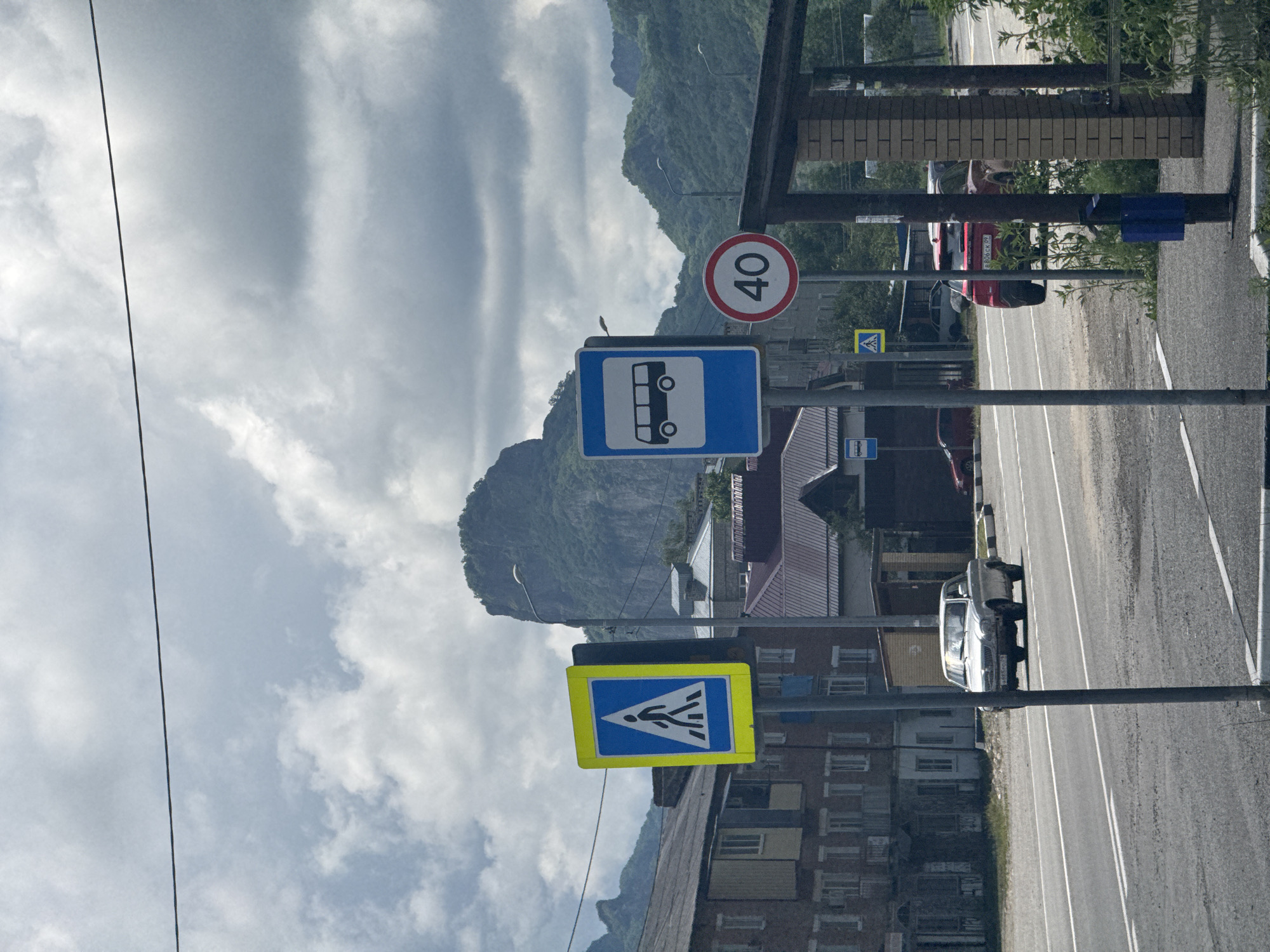 The image shows a street scene in Ordzhonikidzevsky, Russia, with a focus on traffic signs and the surrounding urban and natural landscape. A pedestrian crossing sign, a bus stop sign, and a speed limit sign (40) are prominently displayed. In the background, there are residential buildings and a mountainous terrain. A silver car is driving on the road, and a red car is parked. The sky is partly cloudy.