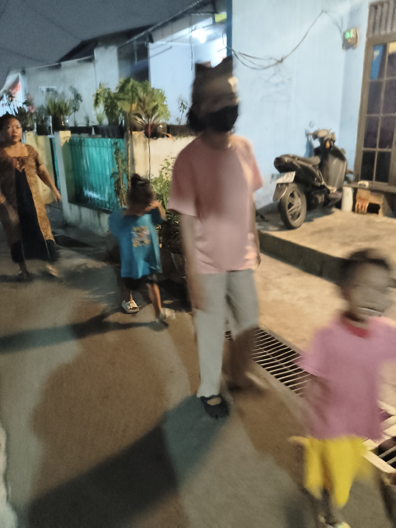 In a street scene at night in South Tangerang, Indonesia, a group of people, including adults and children, are captured in motion. The foreground features a child in a pink shirt and yellow pants, moving from right to left, partially obscured by motion blur. To their left, an adult in a light pink shirt and light-colored pants is also in motion, facing away from the camera. Further to the left, another child in a blue shirt and dark shorts walks with their head down. On the far left, an adult in a patterned brown and black outfit is walking and looking to the left. The environment includes a paved street with a metal grate on the right side, houses with white walls and a tiled roof in the background, and potted plants visible along a green fence. A motorcycle is parked on the right side of the street. The lighting suggests artificial illumination, contributing to the nighttime atmosphere and the motion blur of the subjects.