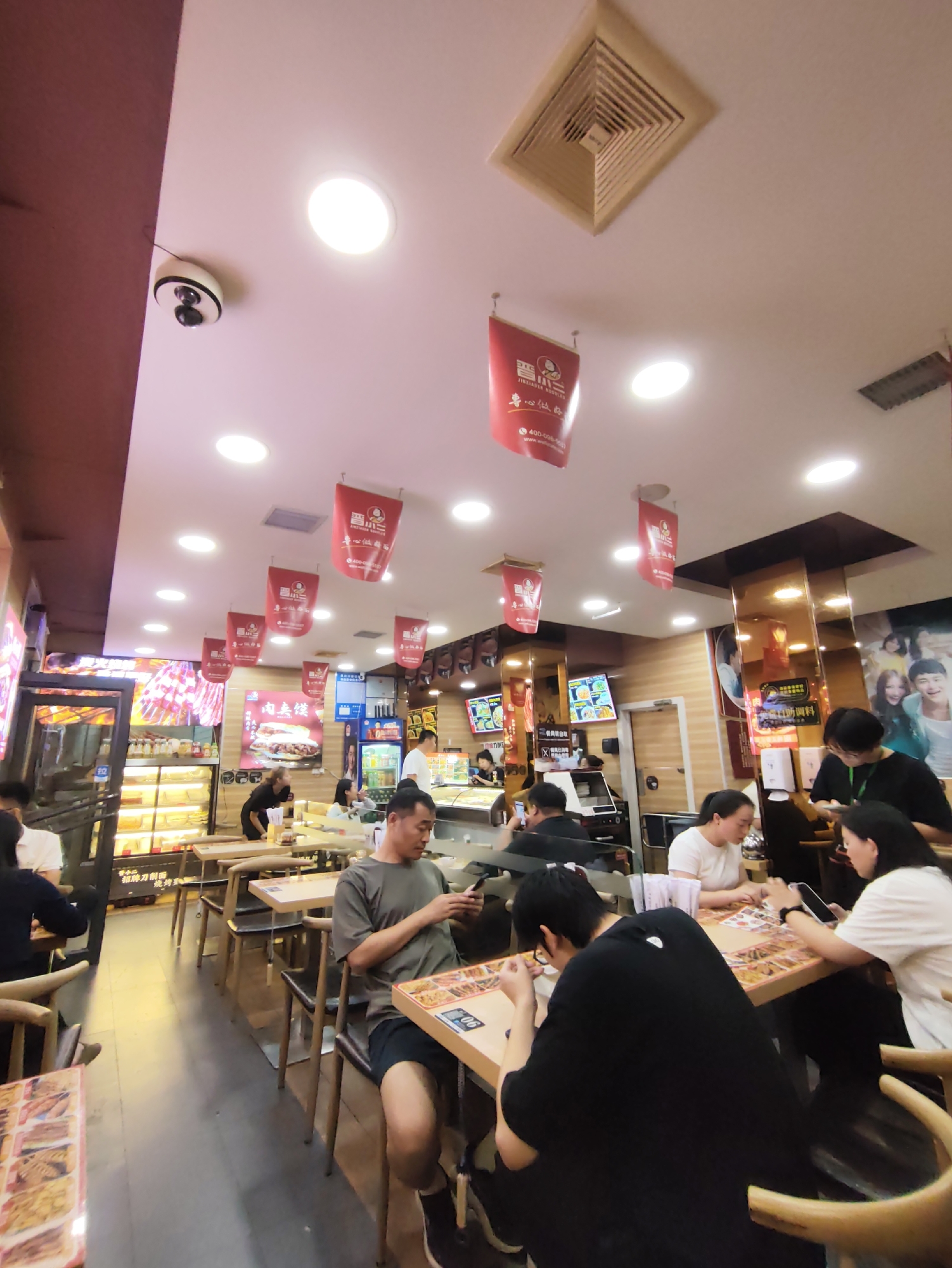The image depicts the interior of a fast-food restaurant in Aihui, China, featuring a bright, overhead lighting system and red banners hanging from the ceiling. Several patrons are seated at wooden tables, some engaged with their mobile phones, others interacting with each other, and one individual is actively eating. The restaurant's service area is visible with a display case containing food items and a counter where staff appear to be preparing orders. The overall ambiance suggests a busy and casual dining environment.