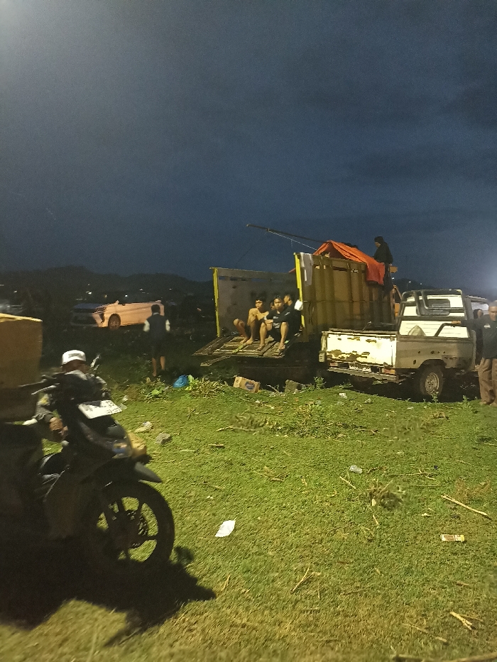 The image captures an evening scene in Kecamatan Pujut, Indonesia, under a dark, cloudy sky, likely at dusk or night. The setting is an open, grassy area, possibly a field, with distant hills visible on the horizon. Artificial lighting, emanating from the right, strongly illuminates the foreground, creating distinct shadows.
In the immediate foreground, a person wearing a white cap is seated on a dark motorcycle. To the right, a box truck with yellow or brownish sides has its tailgate lowered, where three to four young men, some shirtless, are seated. An orange tarp covers part of the truck's cargo, and another person is standing on top of the truck, possibly adjusting the tarp.
Adjacent to the box truck is an older model white pickup truck. A person is standing near the rear of this pickup. Further in the background, to the left, a light-colored sedan or small car is visible with its lights on, appearing to be in a low or uneven patch of ground. Another person stands near this car.
The activity appears to be a casual gathering or a stop, with individuals congregating around the vehicles. The ground is covered in green grass with some scattered debris and boxes. No distinct emotions are discernible, but the scene appears relaxed. There is no clearly legible text other than a partially obscured license plate on the motorcycle.
