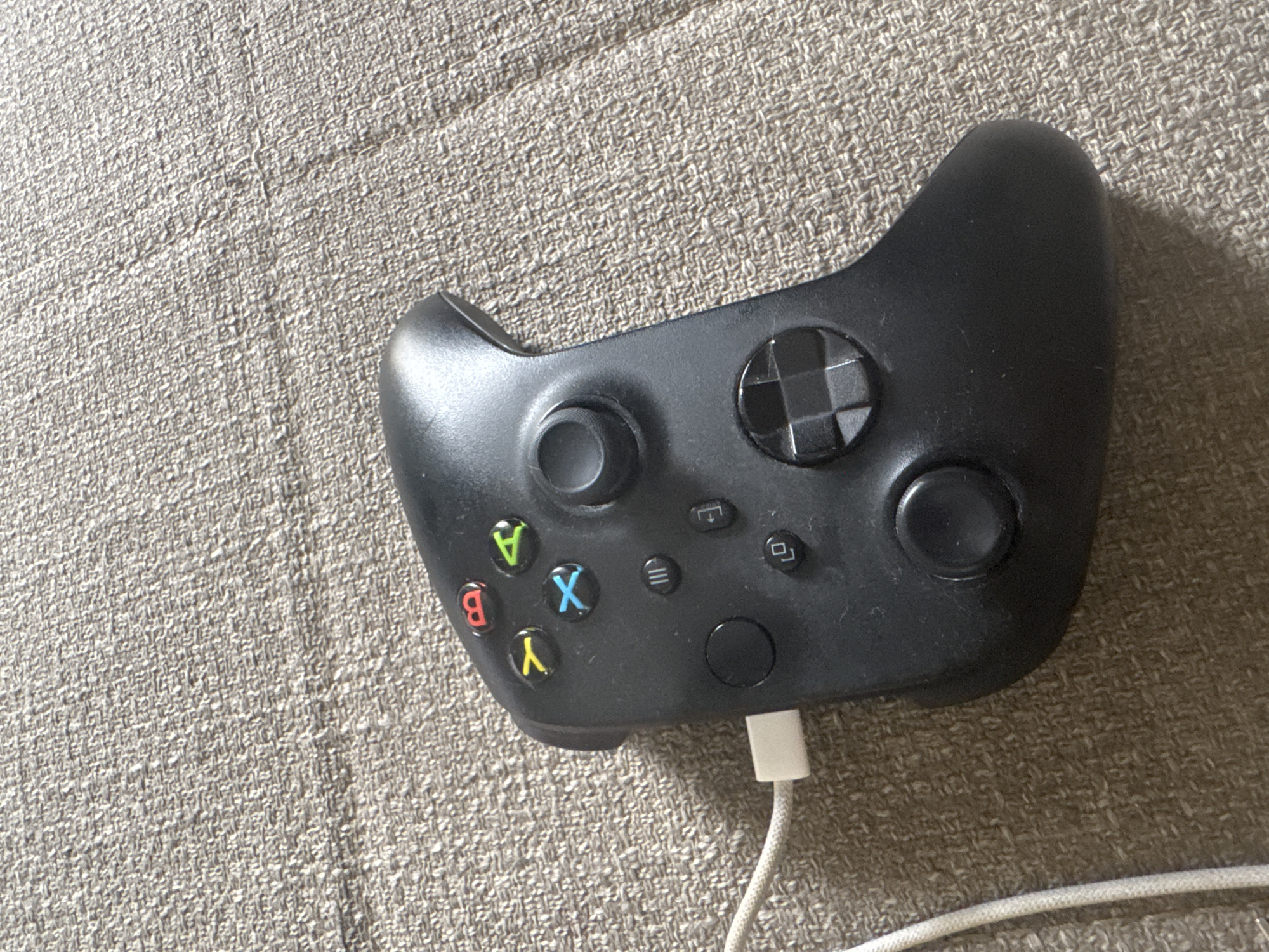 A black Xbox controller is resting on a textured beige surface, likely a sofa or carpet. A white USB-C cable is plugged into the top left of the controller. The controller features the standard button layout, including colored face buttons (A, B, X, Y), a directional pad, analog sticks, and other system buttons. The lighting suggests an indoor setting. There are no people or other objects visible in the frame. The scene does not contain any recognizable landmarks or features that would indicate its location within Tbilisi, Georgia.