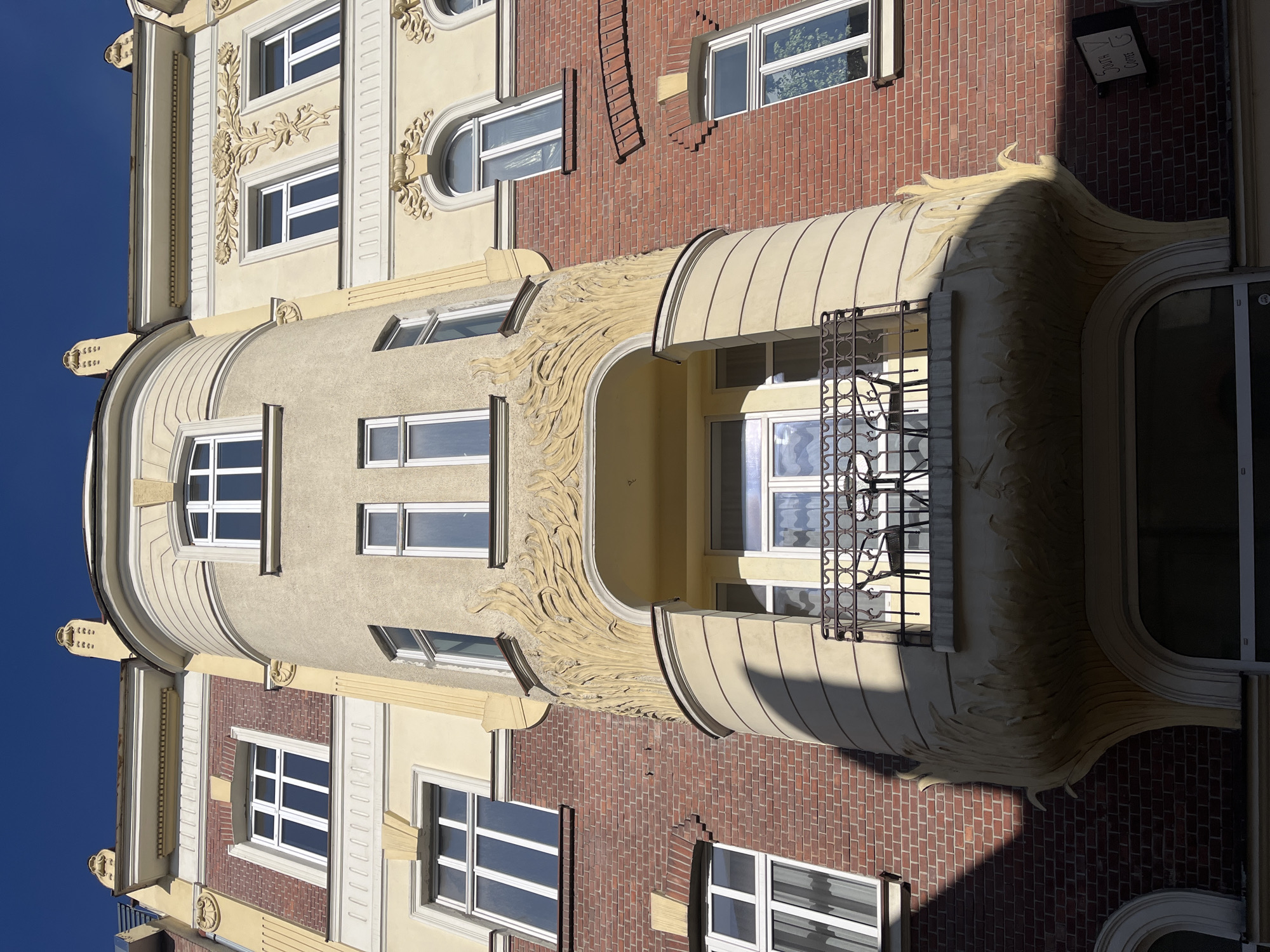 The image shows a building exterior. The building features a cylindrical tower with windows, a balcony with iron railings, and ornate decorations. The facade is a mix of beige, red brick, and decorative floral reliefs. The sky is visible as a clear blue. A sign is partly visible. The building's location is in Unknown, Unknown.