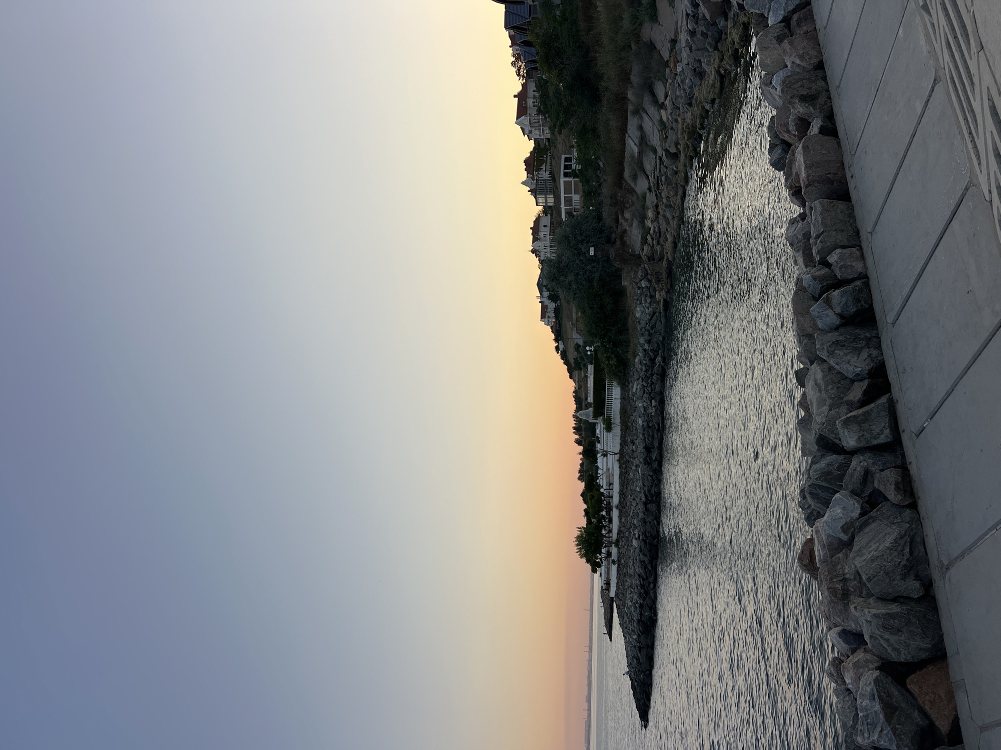 The image shows a tranquil coastal scene at sunset in Odesa, Ukraine. The sky transitions from a clear pale blue at the top to a warm orange and yellow gradient near the horizon. The sea is calm, with gentle ripples reflecting the soft light.
Along the shoreline, a rocky embankment forms a natural barrier, leading into the water. Beyond the rocks, a lush green hillside rises, dotted with several elegant houses, many with distinctive red-tiled roofs. Some of the houses are modern, while others appear to be older, more traditional villas. Trees and vegetation are scattered across the hillside, adding to the picturesque setting.
In the foreground, a concrete walkway runs along the edge of the rocky shoreline. The walkway is made of large, rectangular concrete slabs. The rocky embankment in the foreground is composed of large, grey and brown boulders.
Further out in the water, a small pier or jetty extends from the rocky shore, and a few distant structures are visible on the horizon, possibly part of the city's industrial area. A lone figure can be seen sitting on the rocks near the water's edge, appearing to observe the sunset.
The overall impression is one of peace and natural beauty, with the upscale residential area providing a touch of human habitation against the backdrop of the serene seascape and the fading light of day.